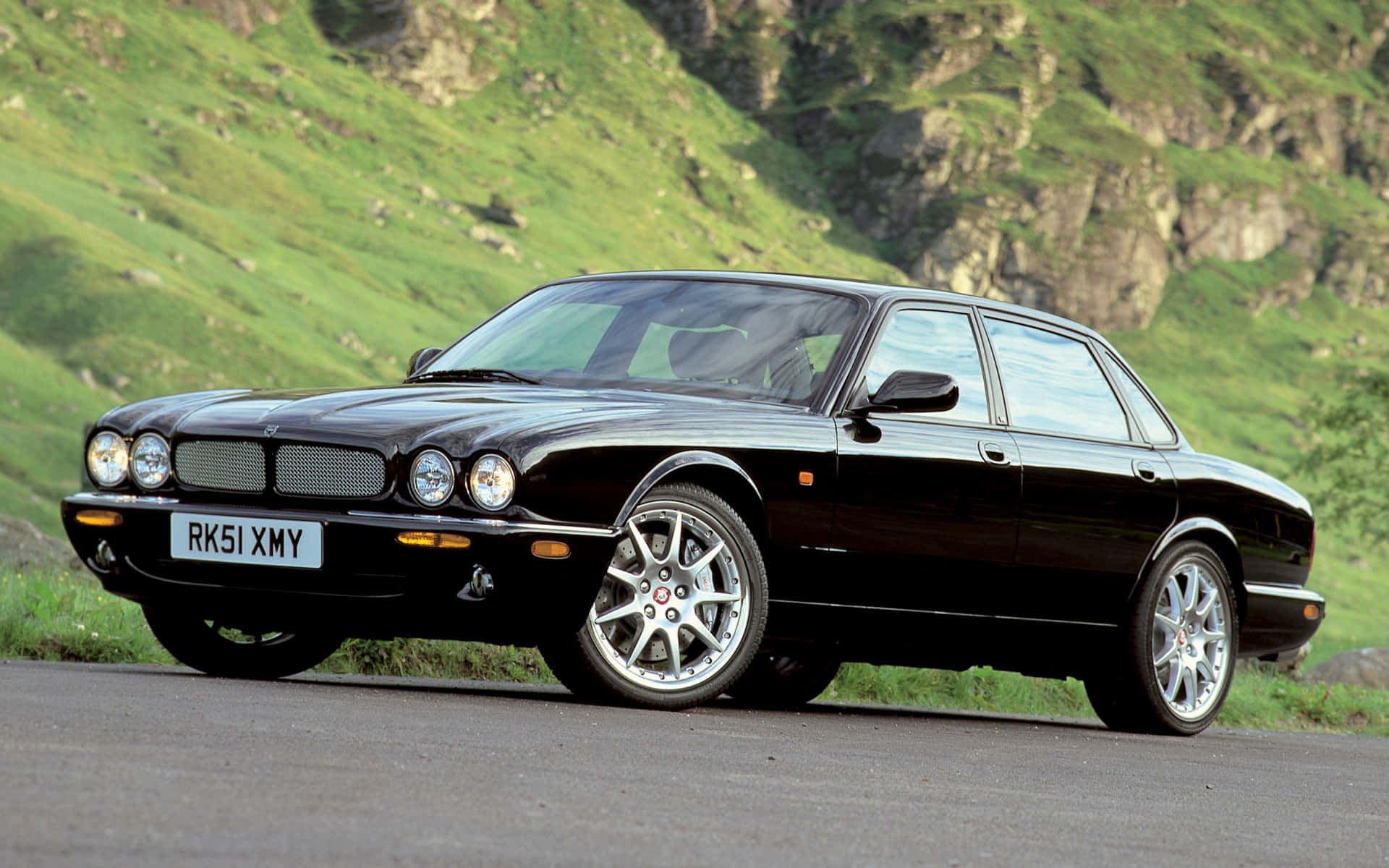 Jaguar Xjr 桌布 壁紙 — 一輛流線型黑色積架XJ轎車沿著蜿蜒的山路行駛，背景是鬱鬱蔥蔥的綠色山丘和岩石峭壁。
