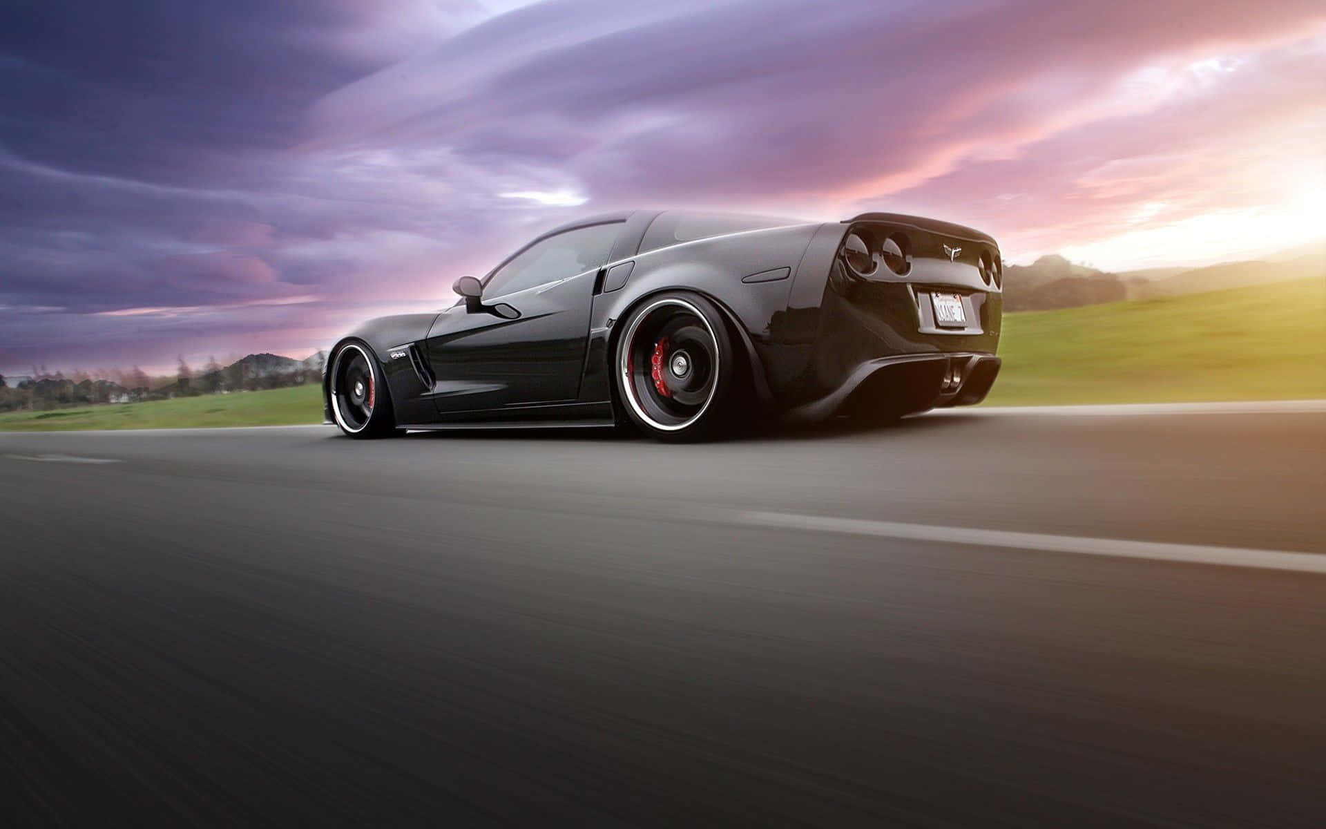 Chevrolet Corvette Z06 桌布 壁紙 — 一輛流線型黑色跑車在日落時分沿著道路飛馳，紫色和橙色的天空籠罩著起伏的綠色山丘。