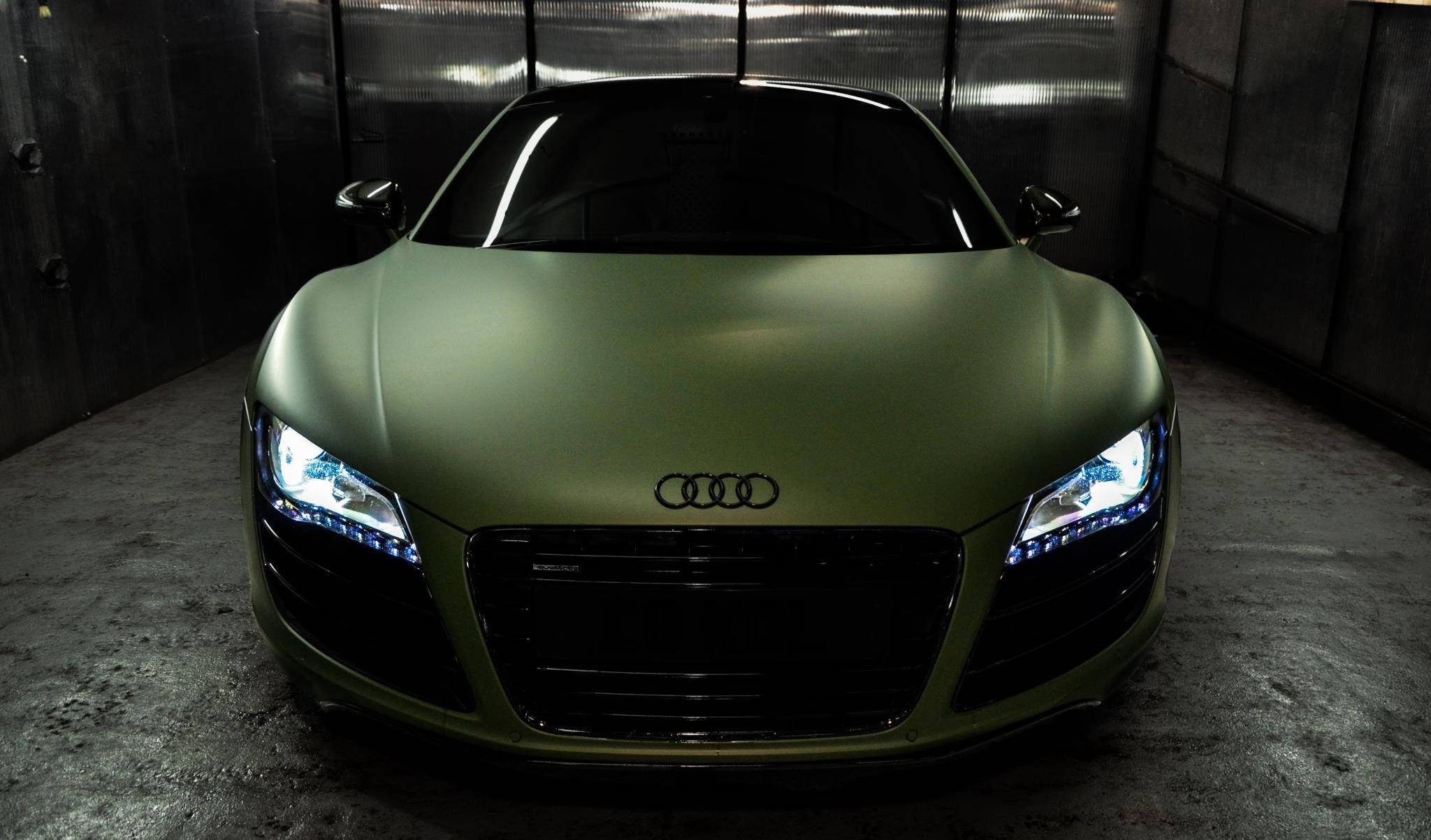 R8 桌布 壁紙 — 一輛消光橄欖綠色的奧迪R8跑車，車頭燈發亮，停放在昏暗的工業車庫中央。