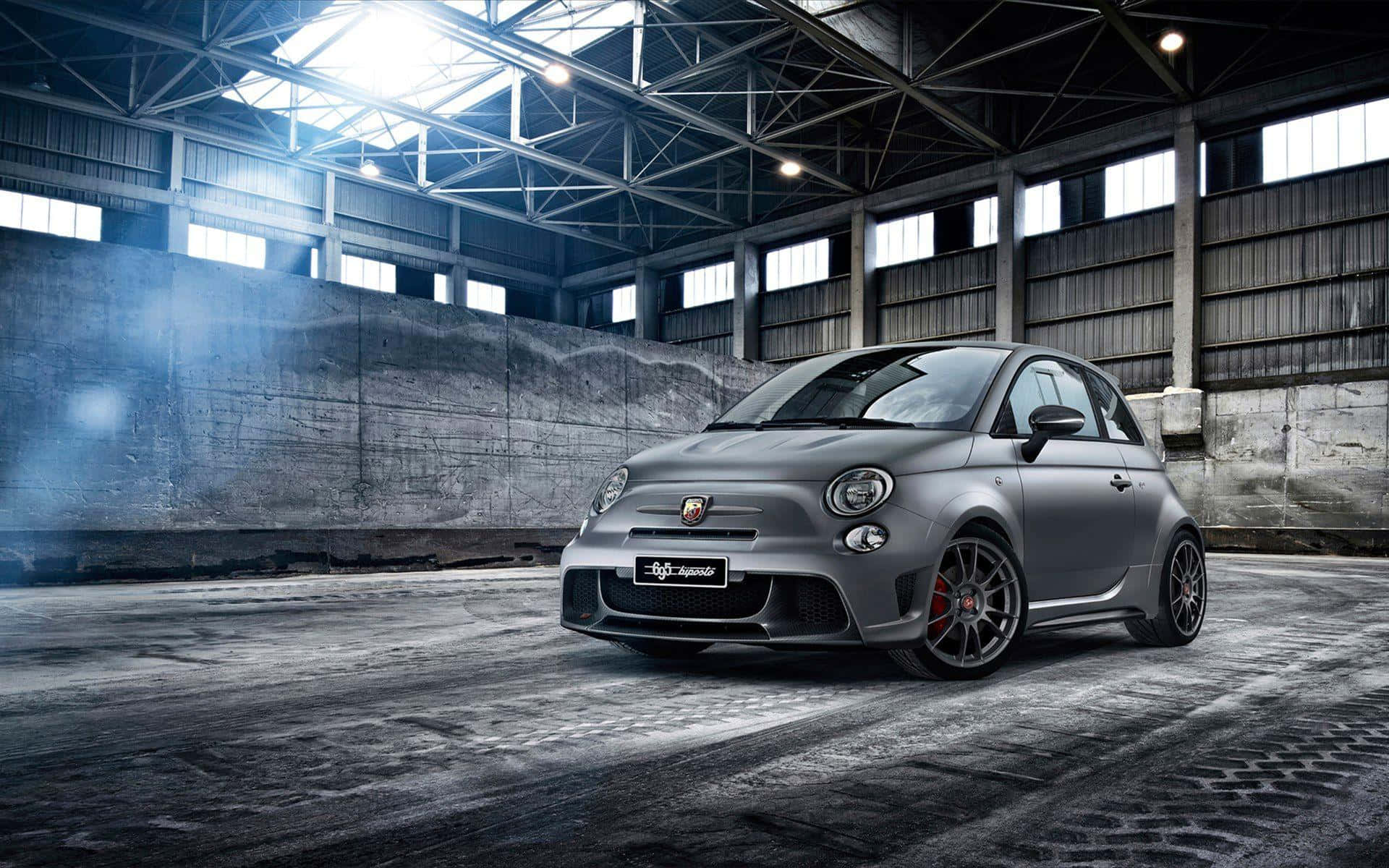 Abarth 桌布 壁紙 — 一輛消光灰色 Fiat 500 Abarth 跑車搭配紅色制動卡鉗，戲劇性地停放在一個充滿灰塵、高聳鋼樑和光線照射的工業倉庫中。