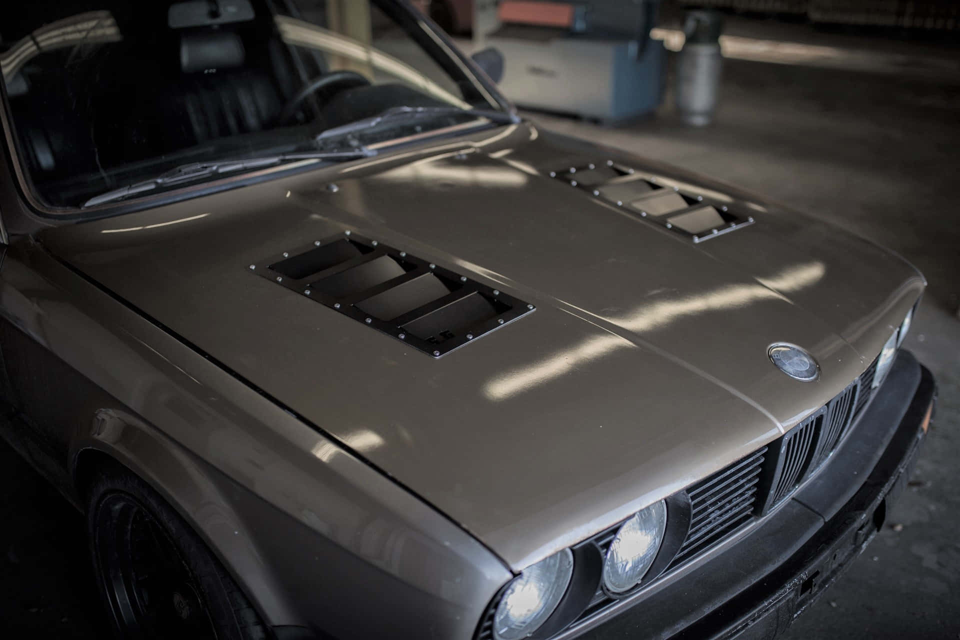 Car Hood 桌布 壁紙 — 一輛消光灰色BMW E30，帶有黑色引擎蓋進氣口，停放在昏暗的車庫中，沐浴在自然光線中。