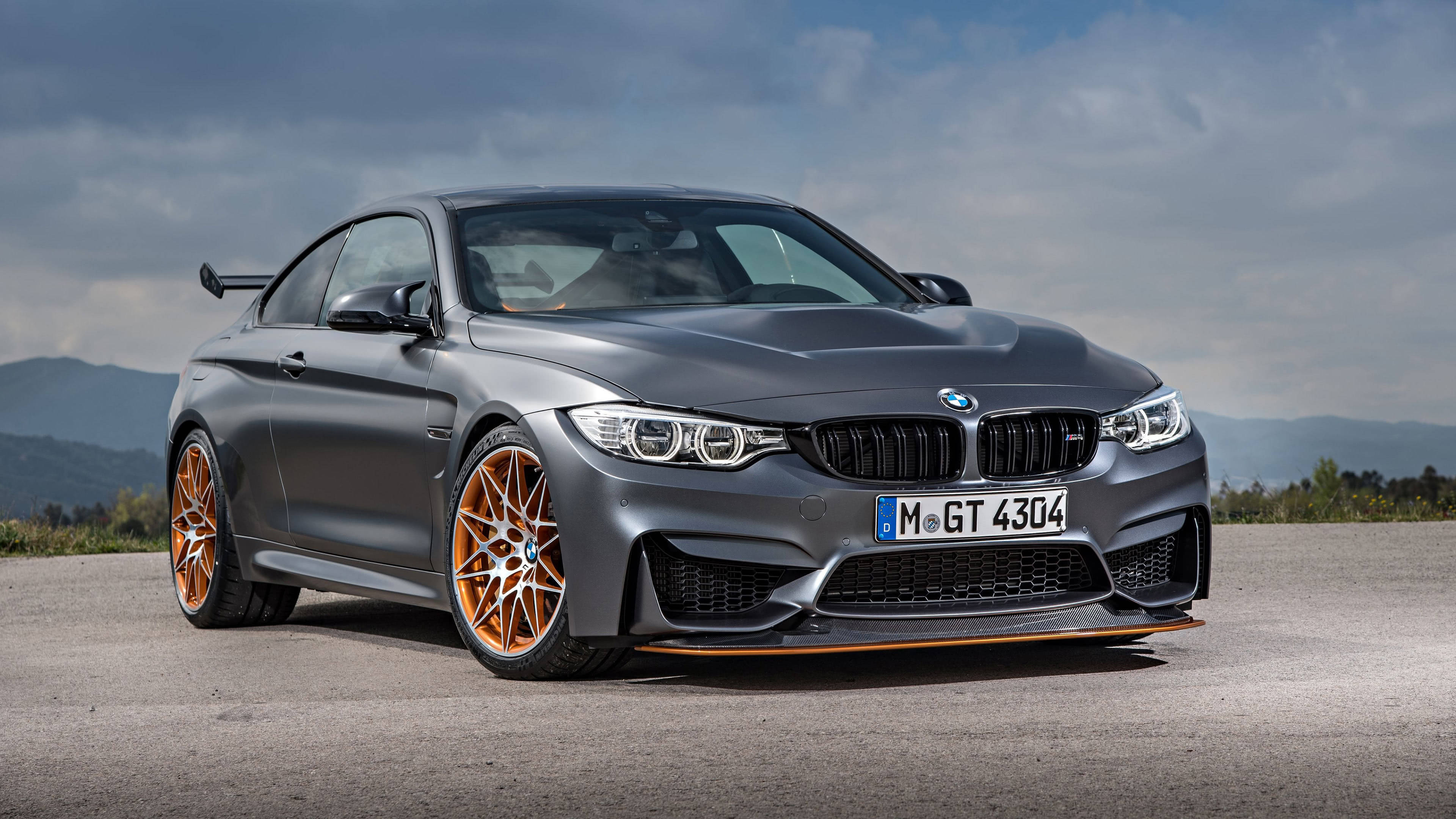 4K BMW 壁紙 桌布 壁紙 — 一輛消光灰色BMW M4 GT4跑車，搭配橘色輪圈，停在柏油路上，背景是陰天和遠山。