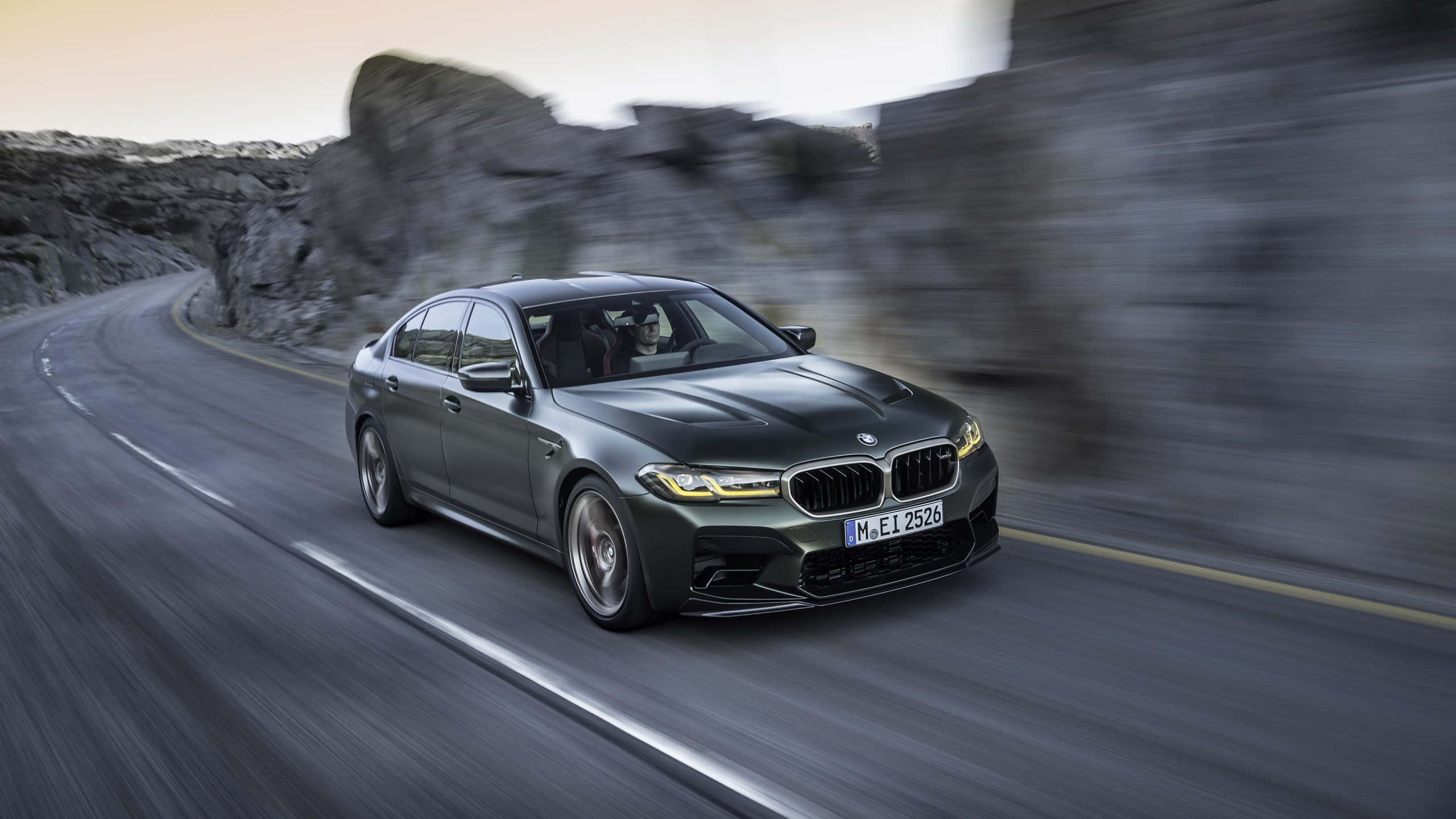 桌布 壁紙 — 一輛消光灰色BMW M5沿著蜿蜒的山路疾駛，伴隨著模糊的岩石懸崖和瀝青動態模糊。