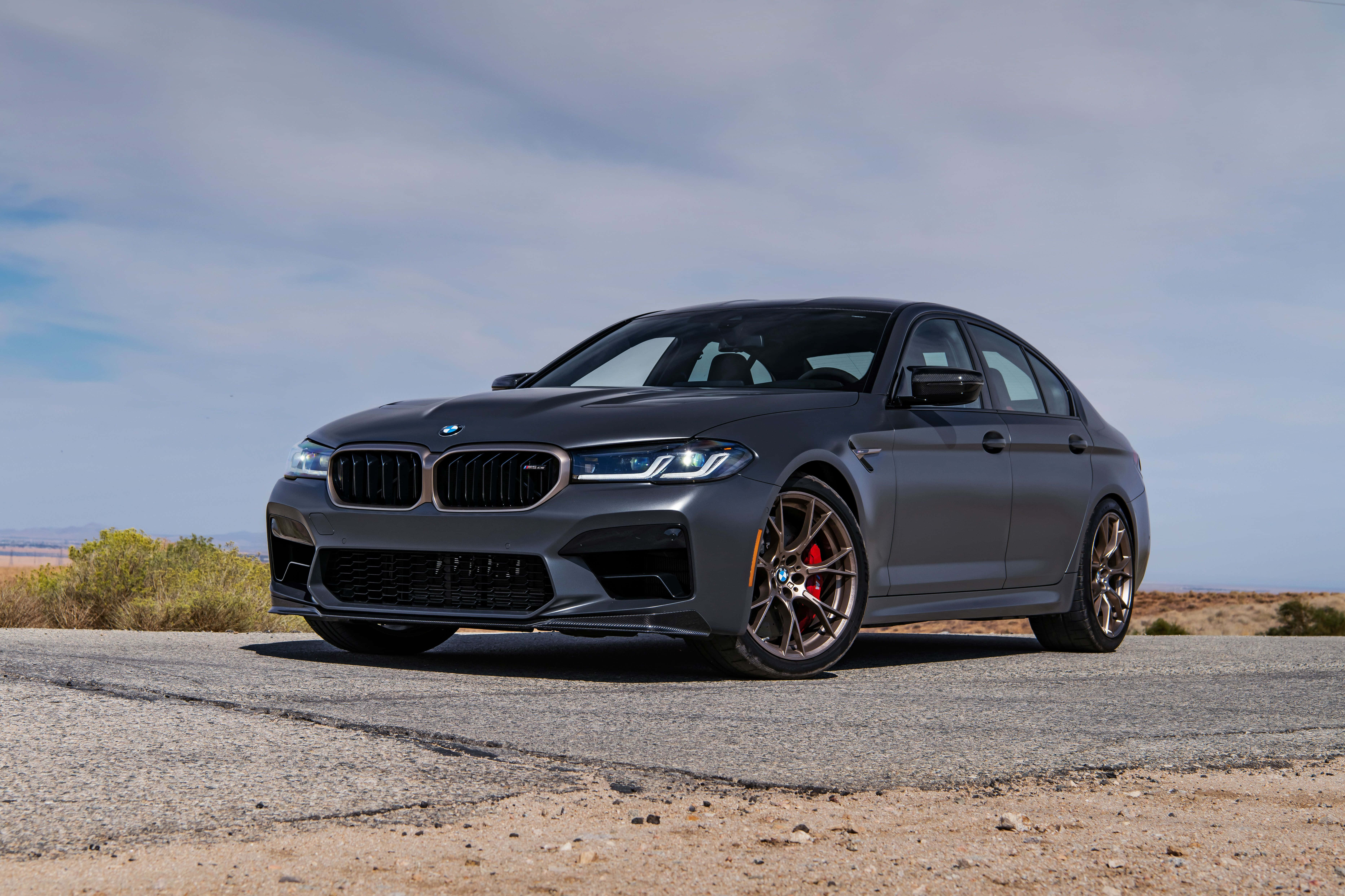 桌布 壁紙 — 一輛消光灰色BMW M5運動轎車，配備青銅輪圈和紅色制動卡鉗，停在乾燥沙漠景觀中一條裂縫的瀝青路上，天空部分多雲。
