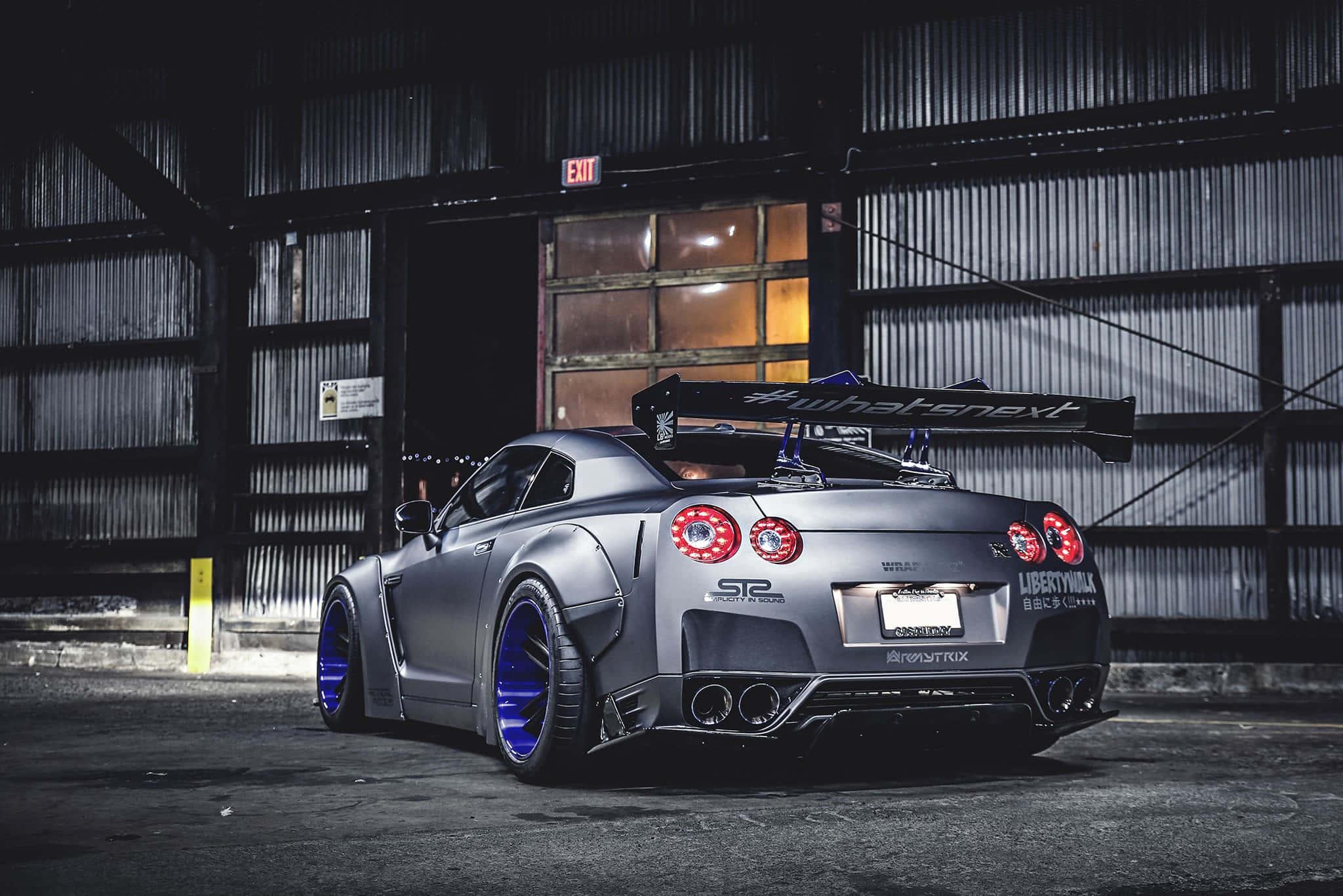 Garage 桌布 壁紙 — 一輛消光灰色日產GT-R，配備藍色輪圈和大型尾翼，停放在燈光昏暗的工業倉庫內。
