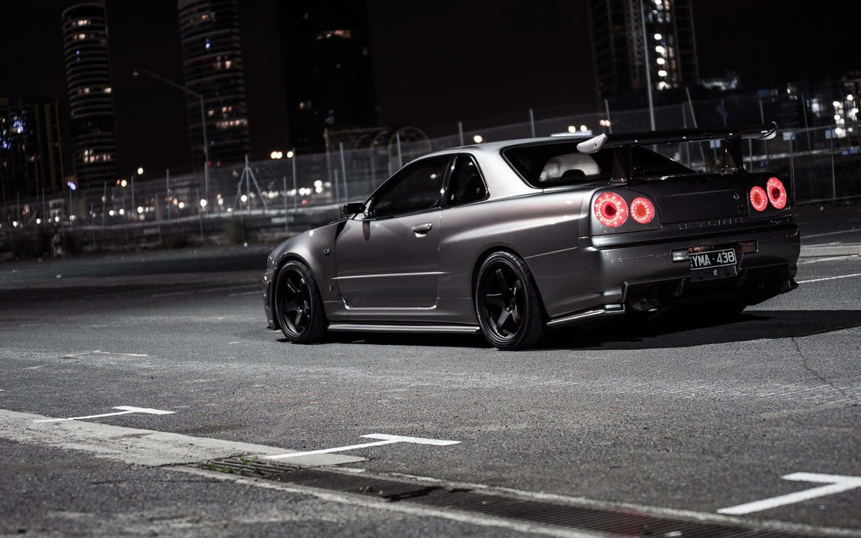 Racing 桌布 壁紙 — 一輛消光灰色日產Skyline GT-R，紅色尾燈發光，夜晚停在摩天大樓背景下的城市街道上。