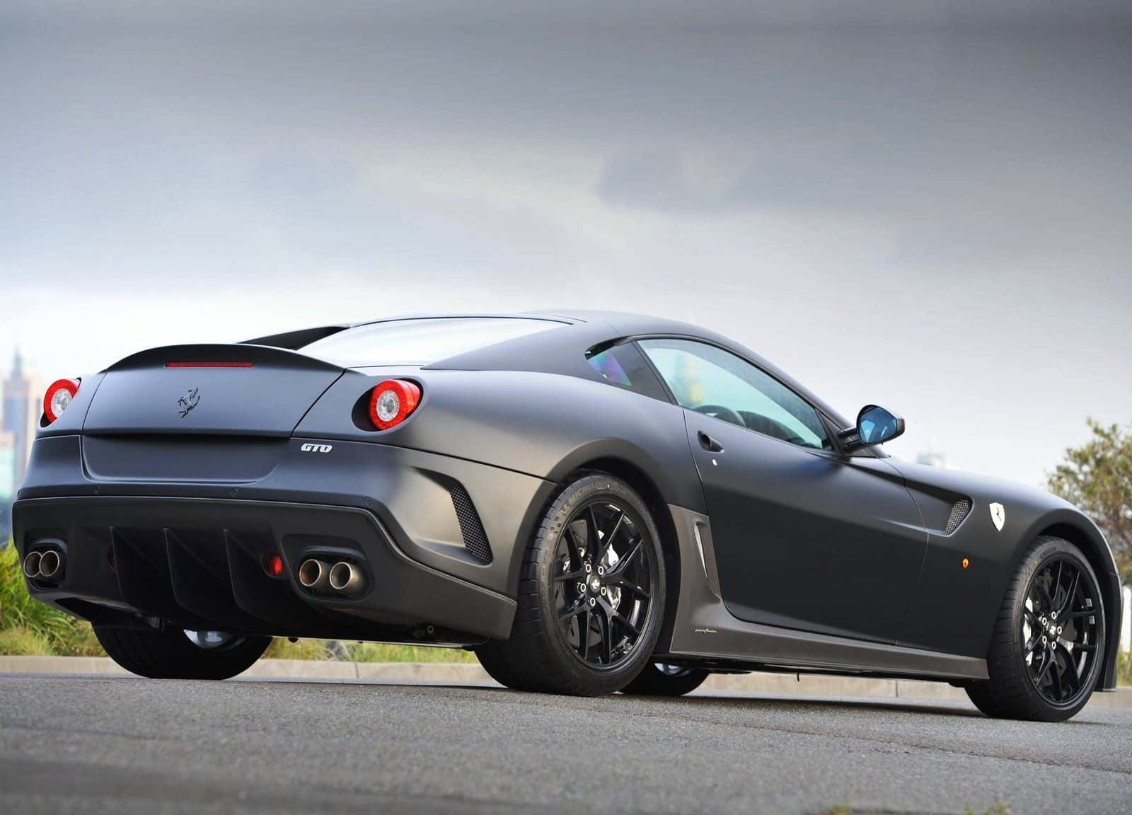 Ferrari 599 Gtb Fiorano 桌布 壁紙 — 一輛消光灰色法拉利 599 GTO 跑車停在柏油路上，天空多雲，遠處有城市景觀。