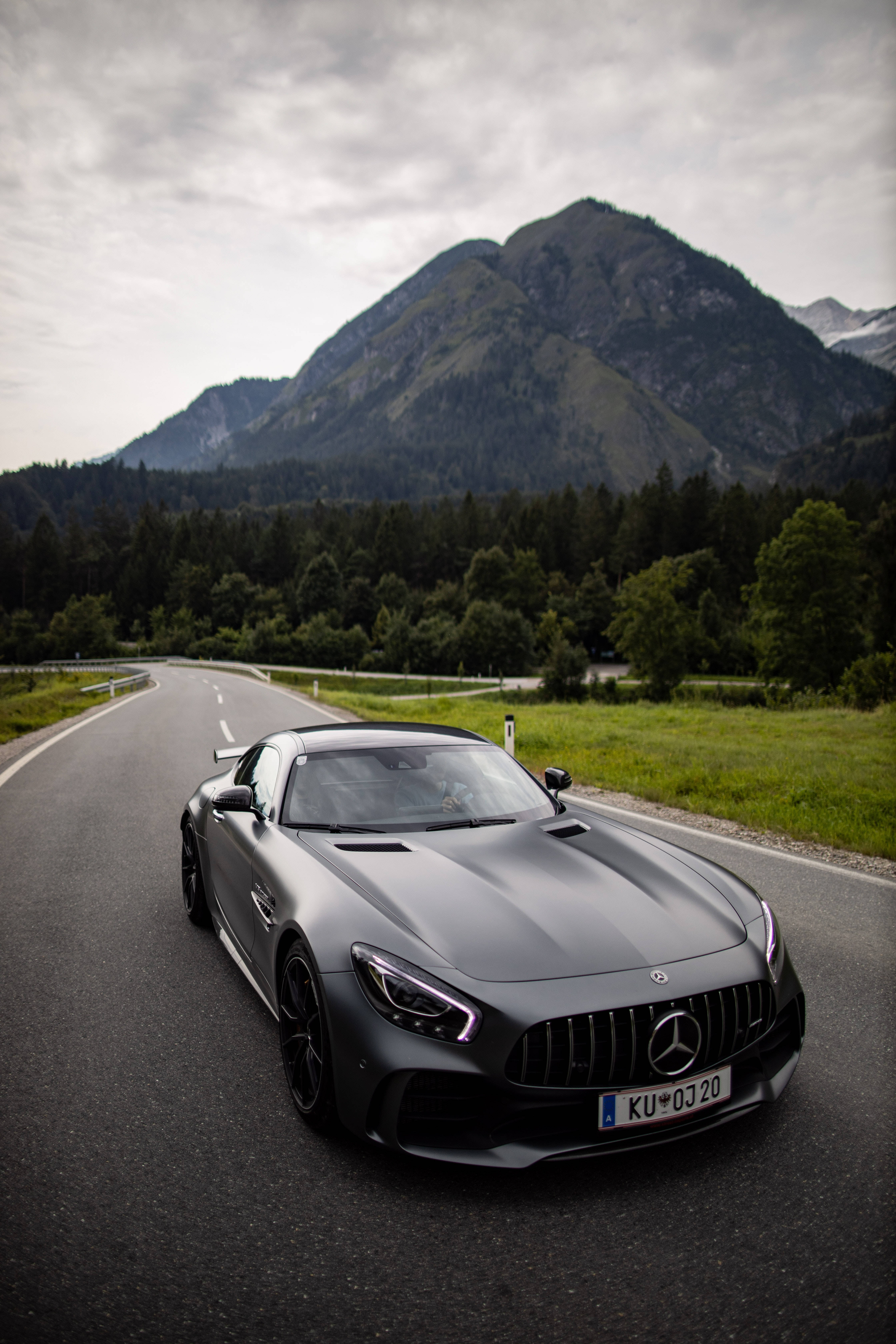 Gtr 桌布 壁紙 — 一輛消光灰色的 Mercedes-AMG GT 跑車沿著蜿蜒的山路行駛，兩旁是茂密的森林和聳立的山峰，天空多雲。