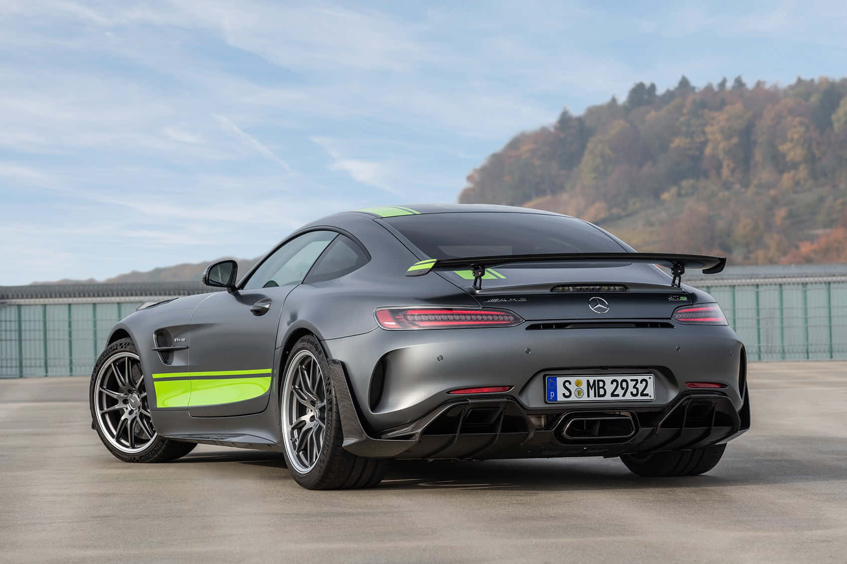 汽車 桌布 壁紙 — 一輛消光灰色的 Mercedes-AMG GT R 跑車，帶有螢光綠色點綴，停放在混凝土表面上，背景是森林覆蓋的山丘，天空部分多雲。