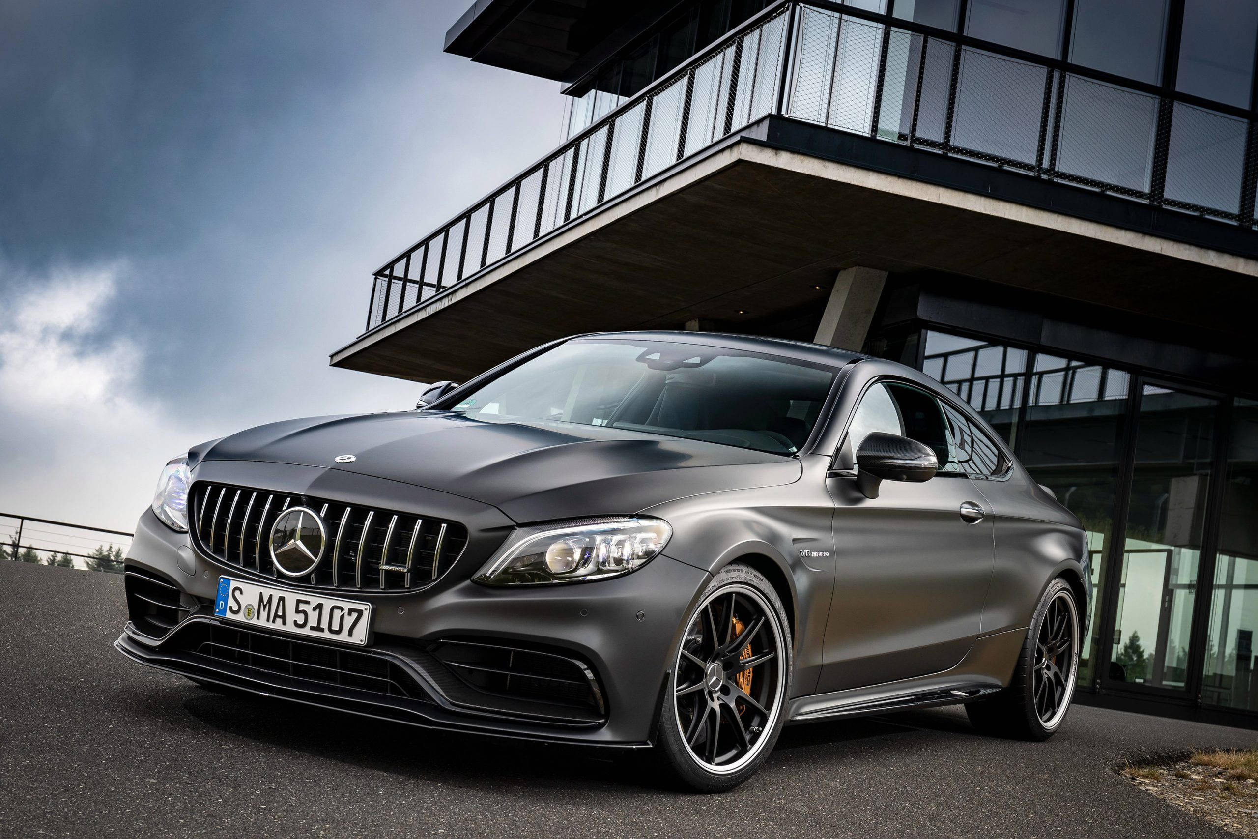 Mercedes Benz Amg 桌布 壁紙 — 一輛消光灰色的 Mercedes-AMG C63 S 雙門轎跑車停放在現代玻璃和鋼結構建築下，天空戲劇性地多雲。