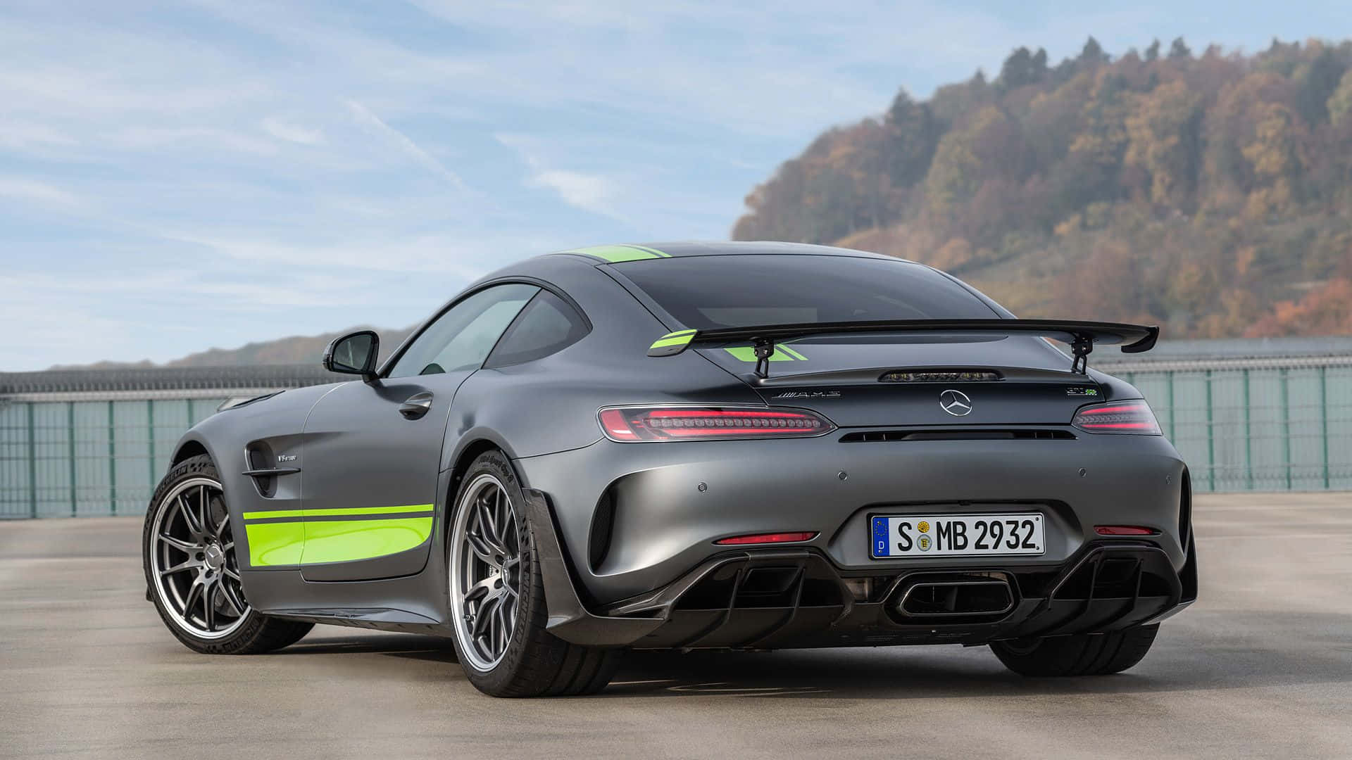 桌布 壁紙 — 一輛消光灰色的 Mercedes-AMG GT R 跑車，帶有螢光綠色點綴，停放在混凝土表面上，背景是晴朗天空下的森林山坡。
