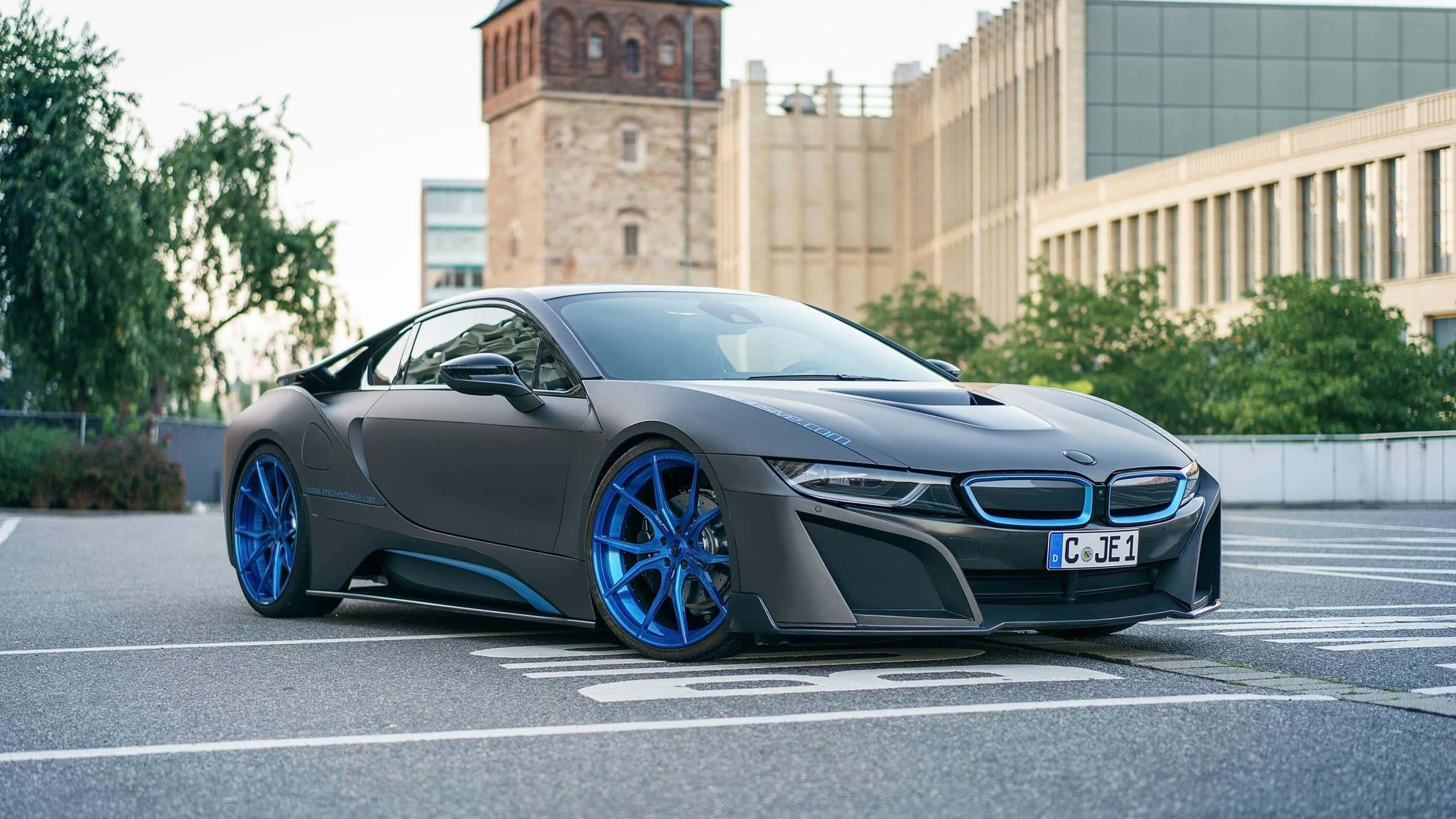 BMW 動圖 桌布 壁紙 — 一輛消光灰色的BMW i8跑車，搭配鮮藍色車輪，停在現代與歷史建築附近的柏油停車場上。
