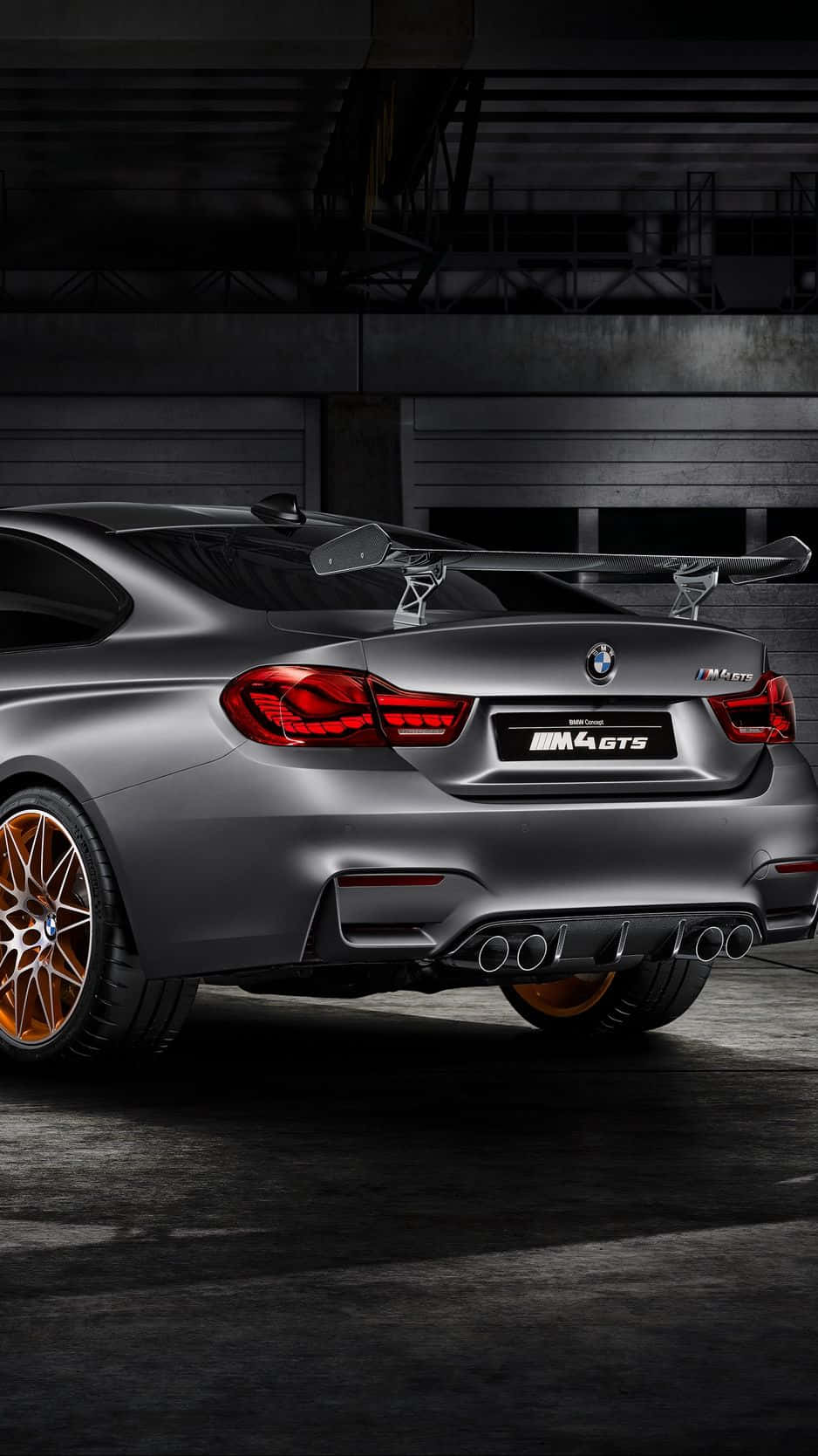Bmw iPhone 桌布 壁紙 — 一輛消光灰色的BMW M4 GTS跑車，配備銅色輪圈和紅色尾燈，停放在昏暗的工業車庫中。