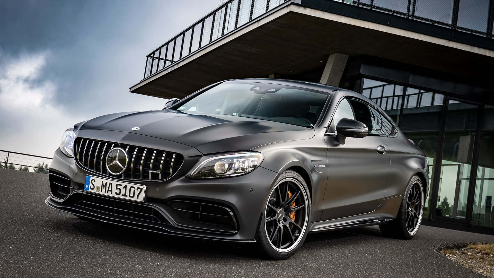 Mercedes 桌布 壁紙 — 一輛消光灰色的Mercedes-AMG C63 S Coupe停放在現代玻璃和鋼結構下，天空戲劇性地多雲。
