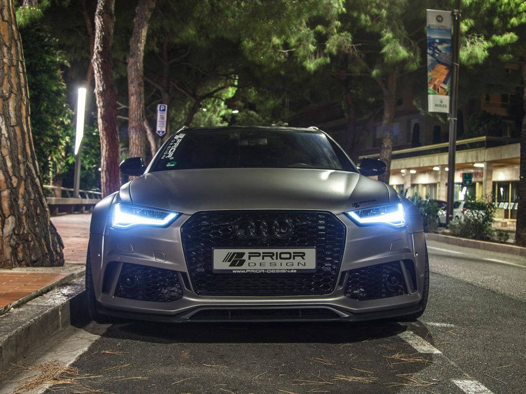 奧迪 桌布 壁紙 — 一輛消光灰色的奧迪RS7，帶有發光的藍色車頭燈，夜間停放在城市街道的松樹下。