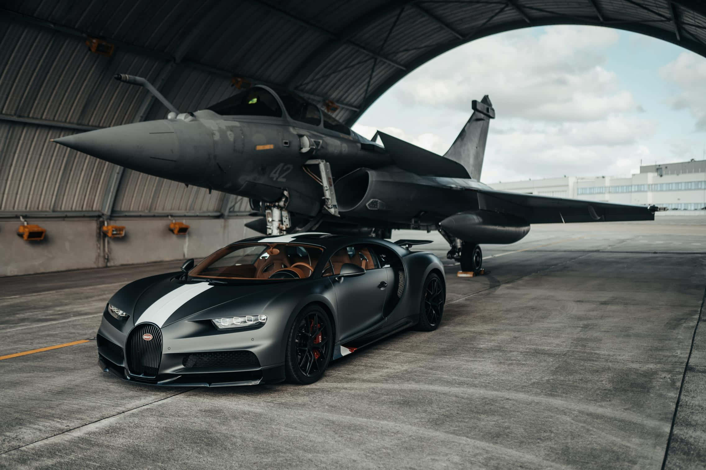 Best Bugatti 桌布 壁紙 — 一輛消光灰色的布加迪Chiron停在一架流線型戰鬥機下方，位於廣闊的工業機庫內，天空多雲。