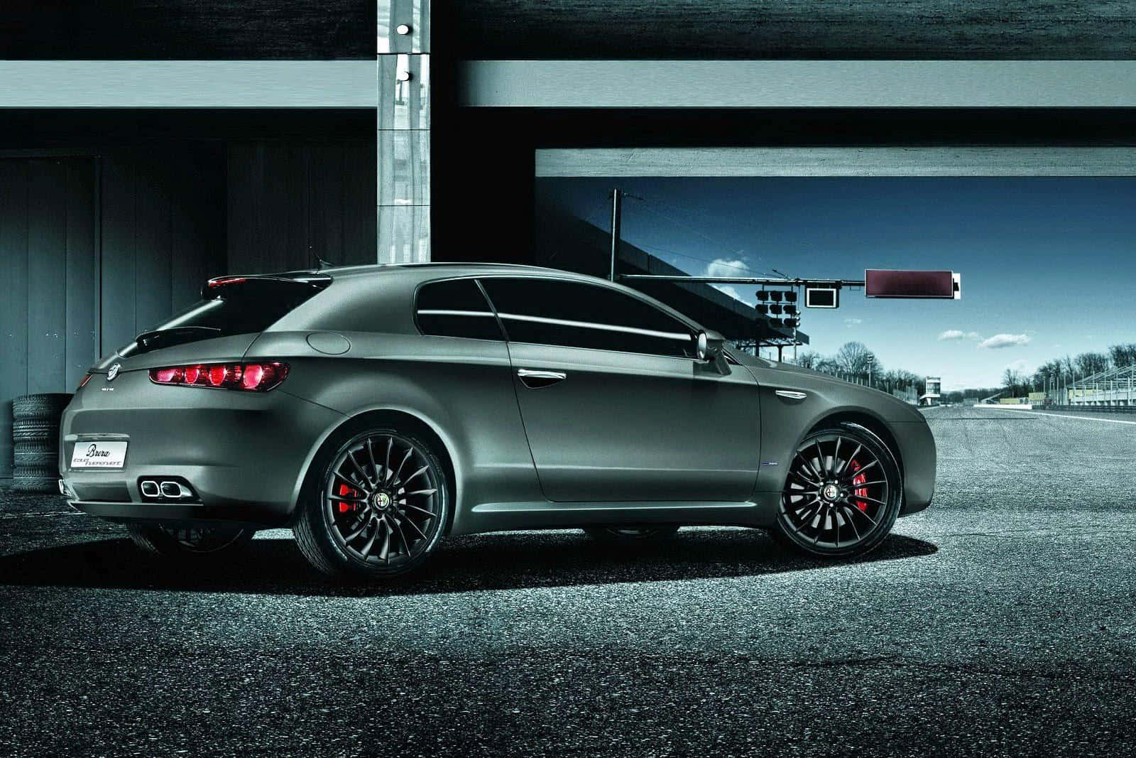 Alfa Romeo Brera 桌布 壁紙 — 一輛消光灰色的愛快羅密歐跑車，配備黑色輪圈和紅色煞車卡鉗，停放在現代結構下的賽道上。