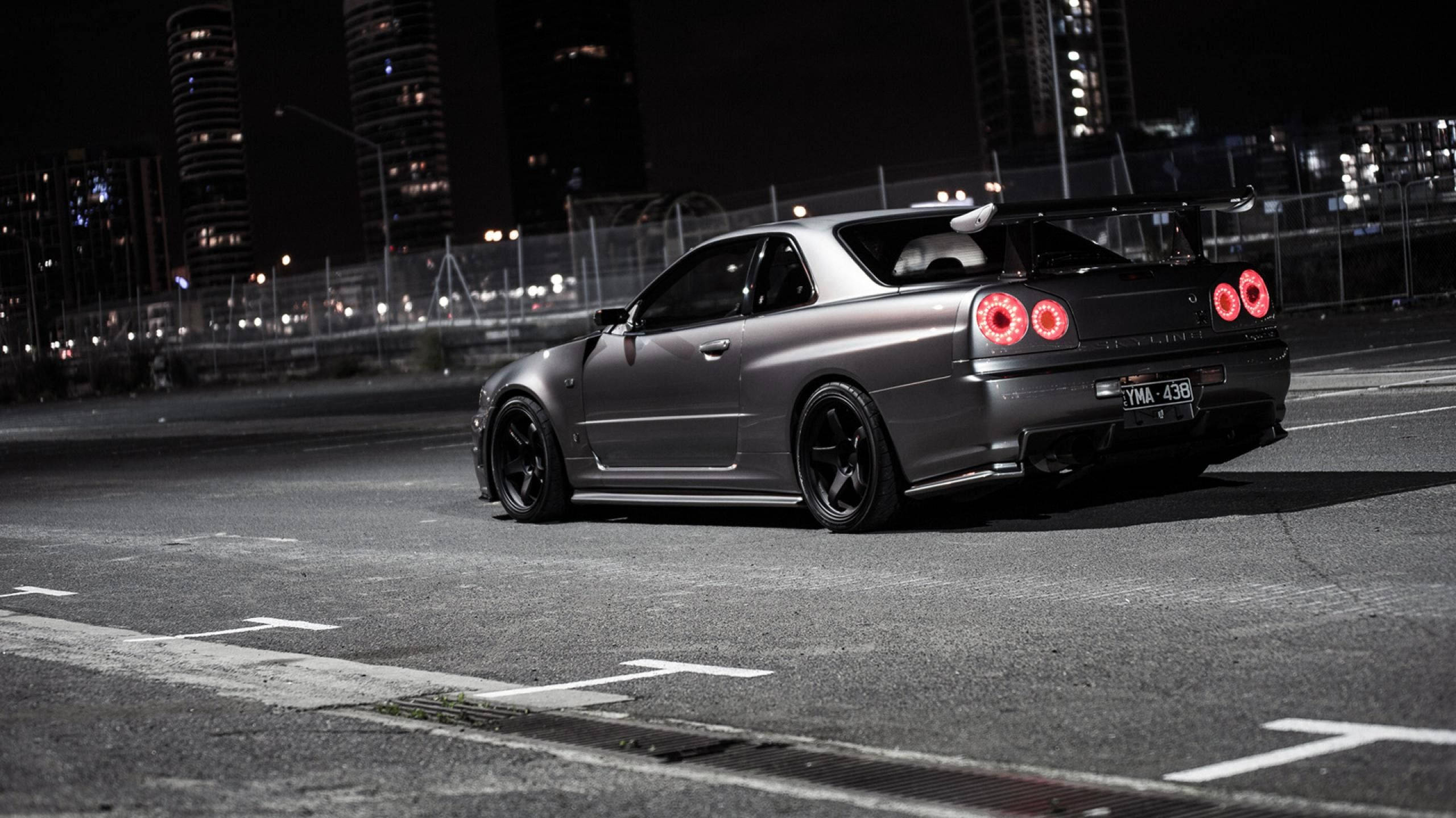 Nissan 桌布 壁紙 — 一輛消光灰色的日產Skyline GT-R停在夜晚的城市街道上，紅色尾燈發光，背景是燈火通明的摩天大樓。