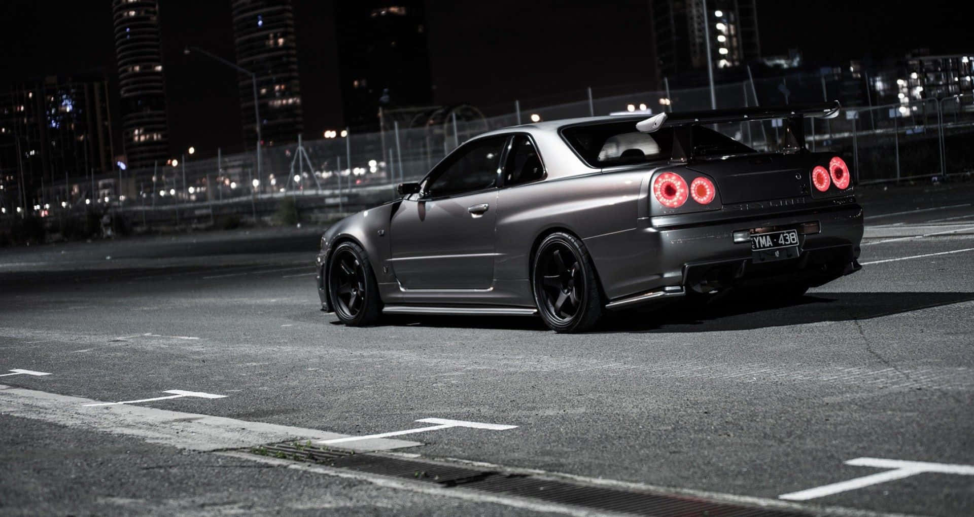 JDM汽車 桌布 壁紙 — 一輛消光灰色的日產Skyline R34跑車，帶有發光的紅色尾燈，夜晚停在空無一人的城市街道上。