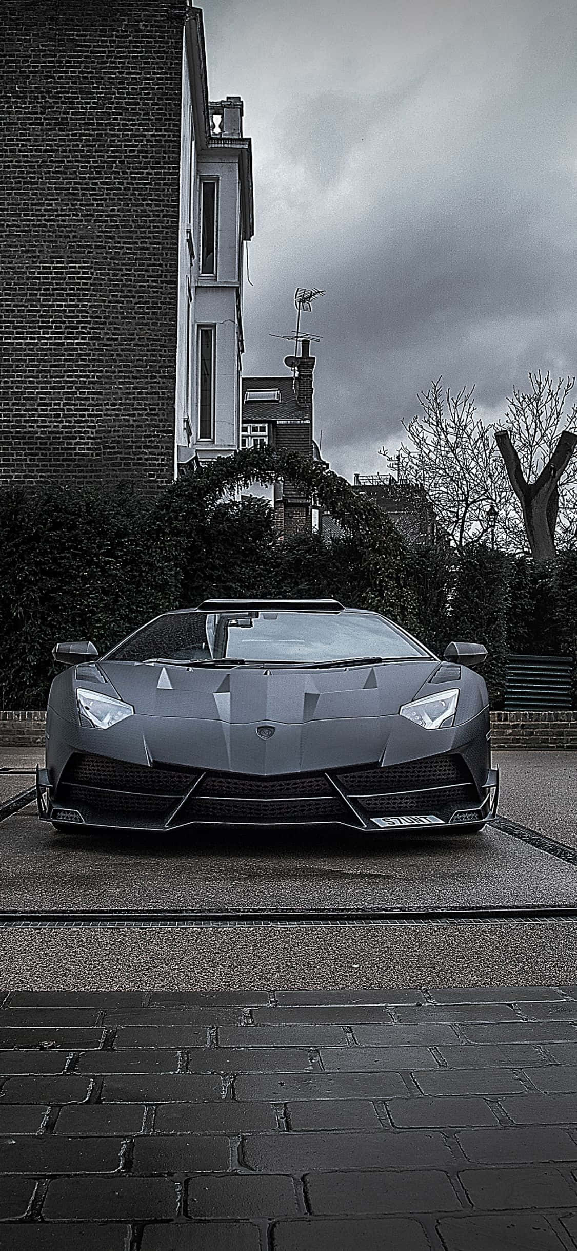Lamborghini Phone 桌布 壁紙 — 一輛消光灰色的藍寶堅尼 Aventador 停泊在濕漉漉的鵝卵石街道上，天空陰沉多雲。