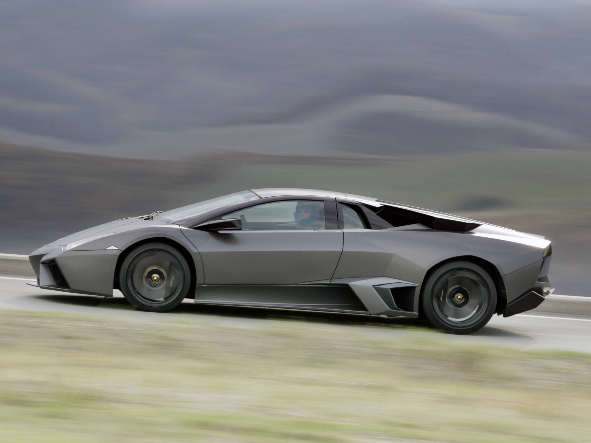4K Lamborghini 桌布 壁紙 — 一輛消光灰色的藍寶堅尼Reventon在蜿蜒的道路上行駛，背景是模糊的山丘。