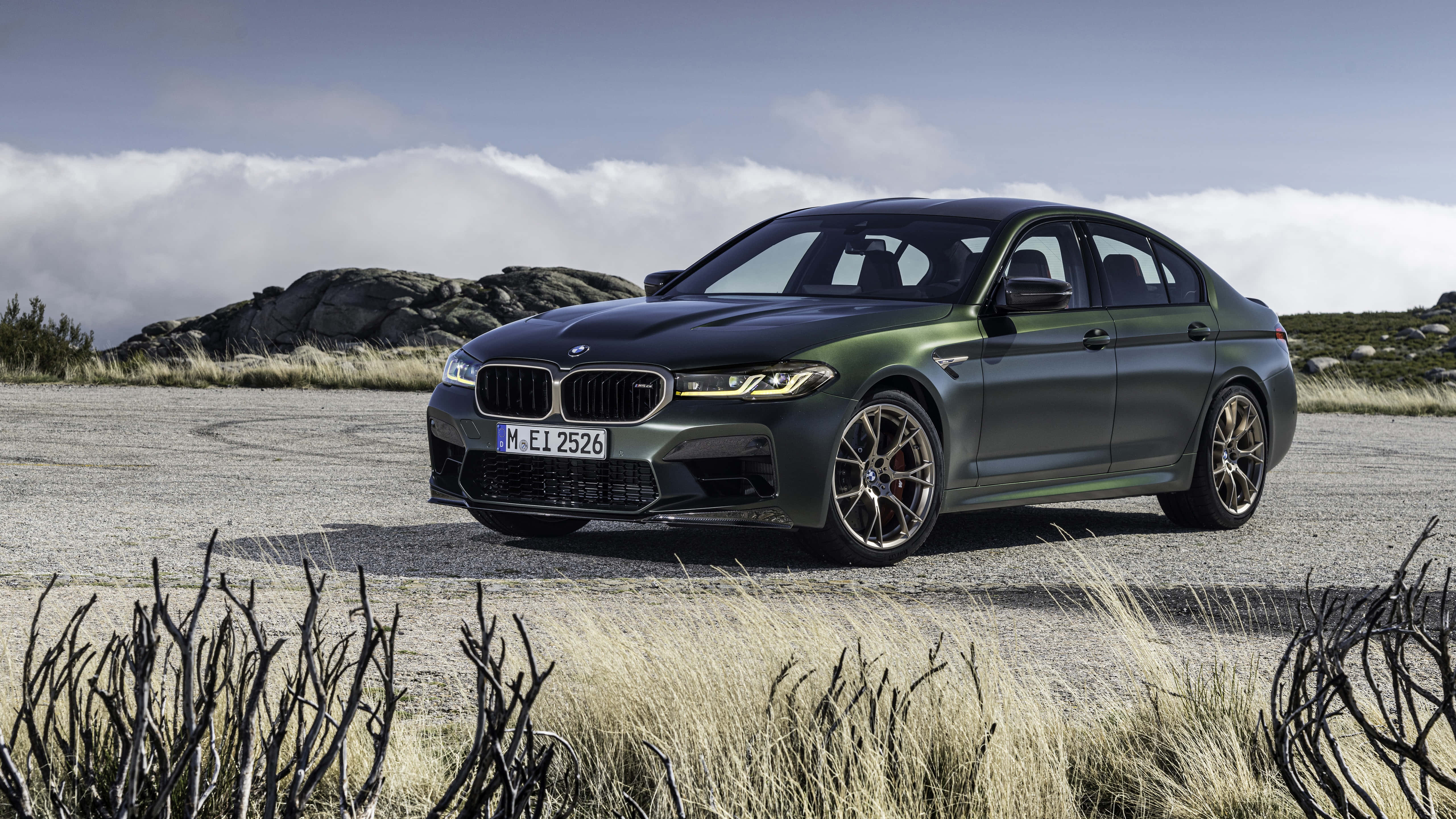 桌布 壁紙 — 一輛消光綠色BMW M5運動轎車停在碎石路上，周圍有岩石山丘和乾草，天空多雲。