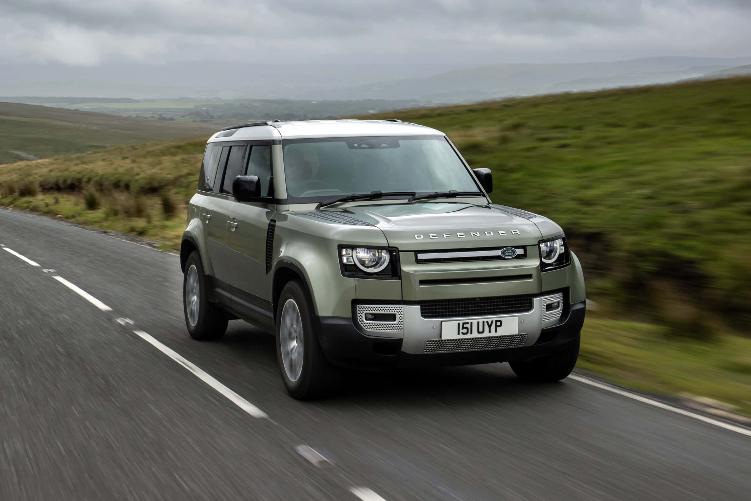 Land Rover Defender 桌布 壁紙 — 一輛消光綠色的 Land Rover Defender 在陰天下沿著蜿蜒的鄉間道路奔馳，背景是模糊的綠色丘陵。