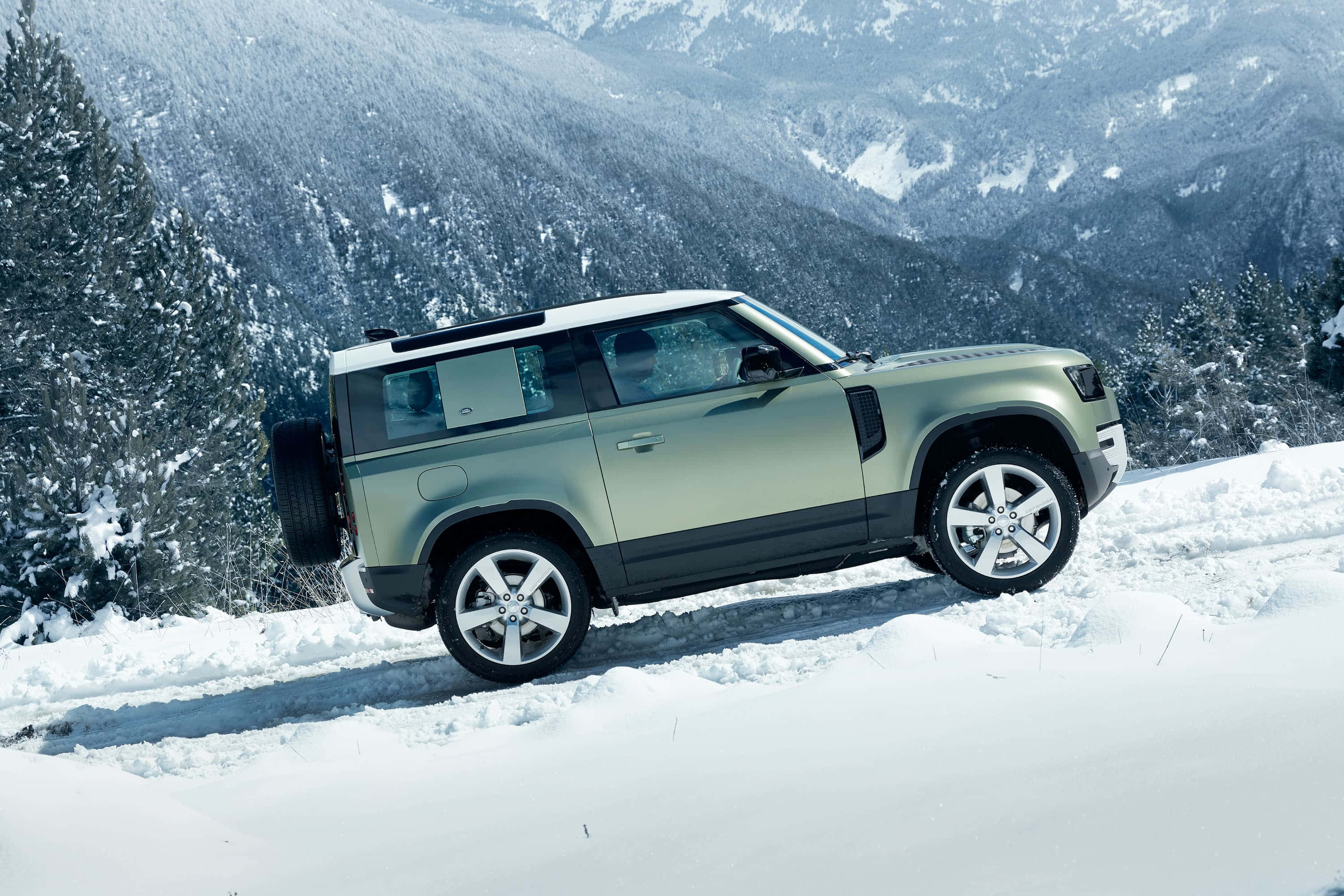 Land Rover Defender 桌布 壁紙 — 一輛消光綠色的Land Rover Defender在積雪深厚的山路上行駛，兩旁是白雪覆蓋的松樹。