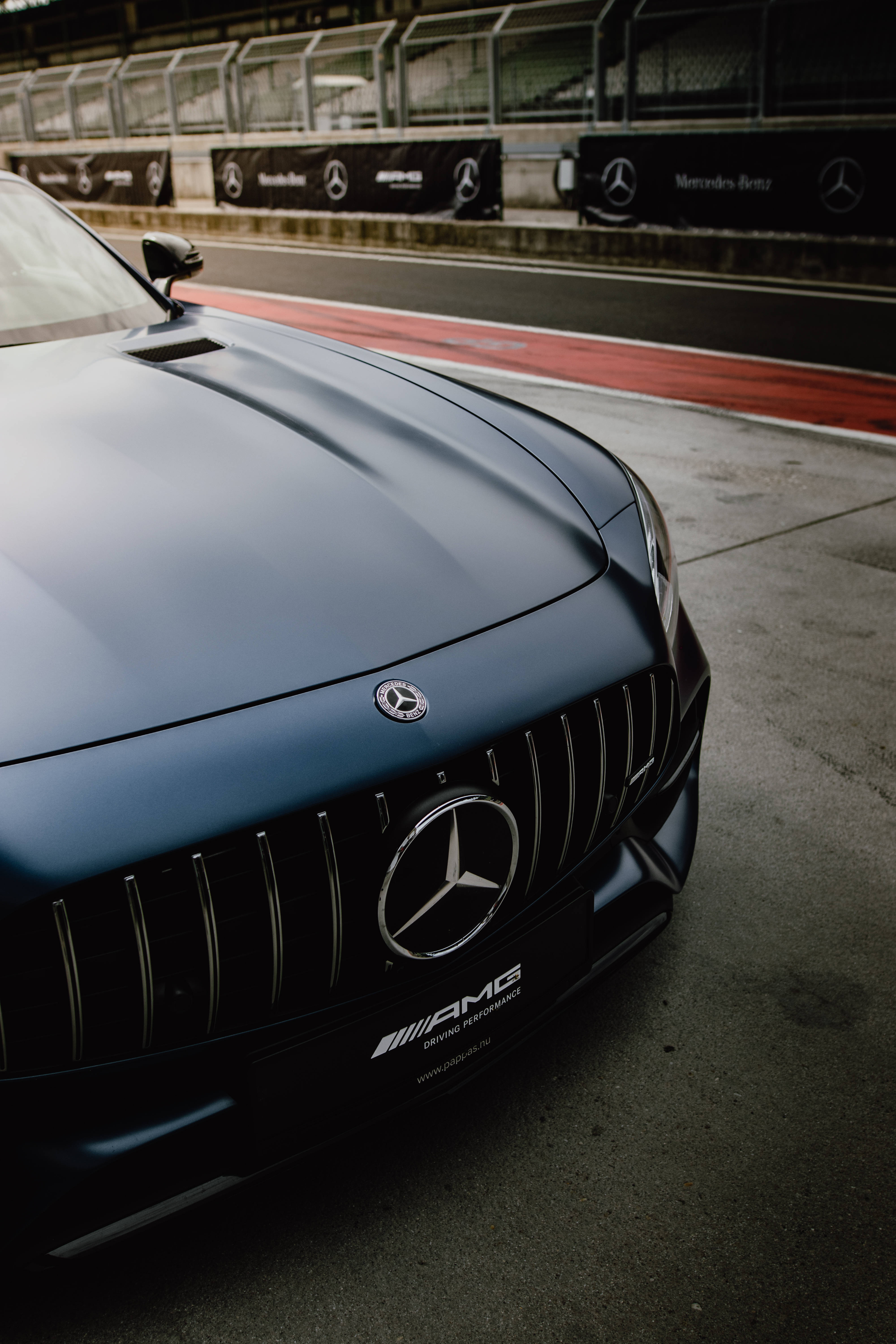 amg iPhone 桌布 壁紙 — 一輛消光藍色 Mercedes-AMG 跑車停在賽車場上，背景有紅色路緣和品牌障礙物。