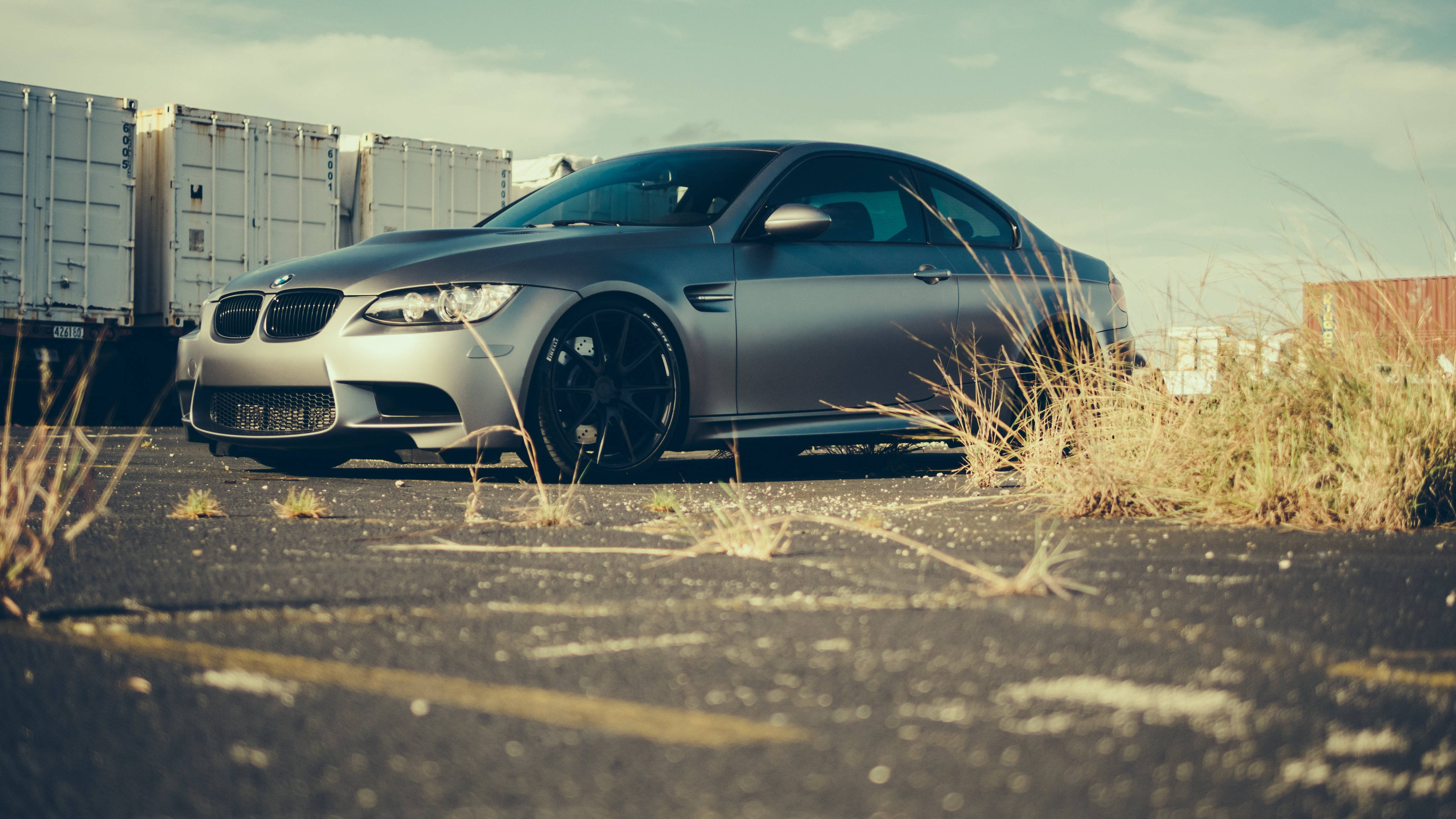 m 桌布 壁紙 — 一輛消光銀色BMW M3雙門跑車停在裂開的柏油路上，背景有乾草和貨運貨櫃，天空朦朧。