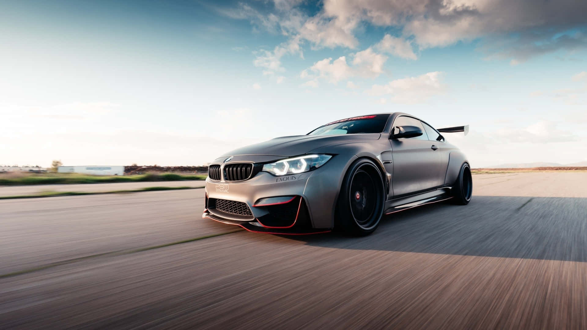 1920x1080 Bmw 桌布 壁紙 — 一輛消光銀色BMW M4雙門跑車帶有紅色點綴，在部分多雲的天空下沿沙漠跑道奔馳。
