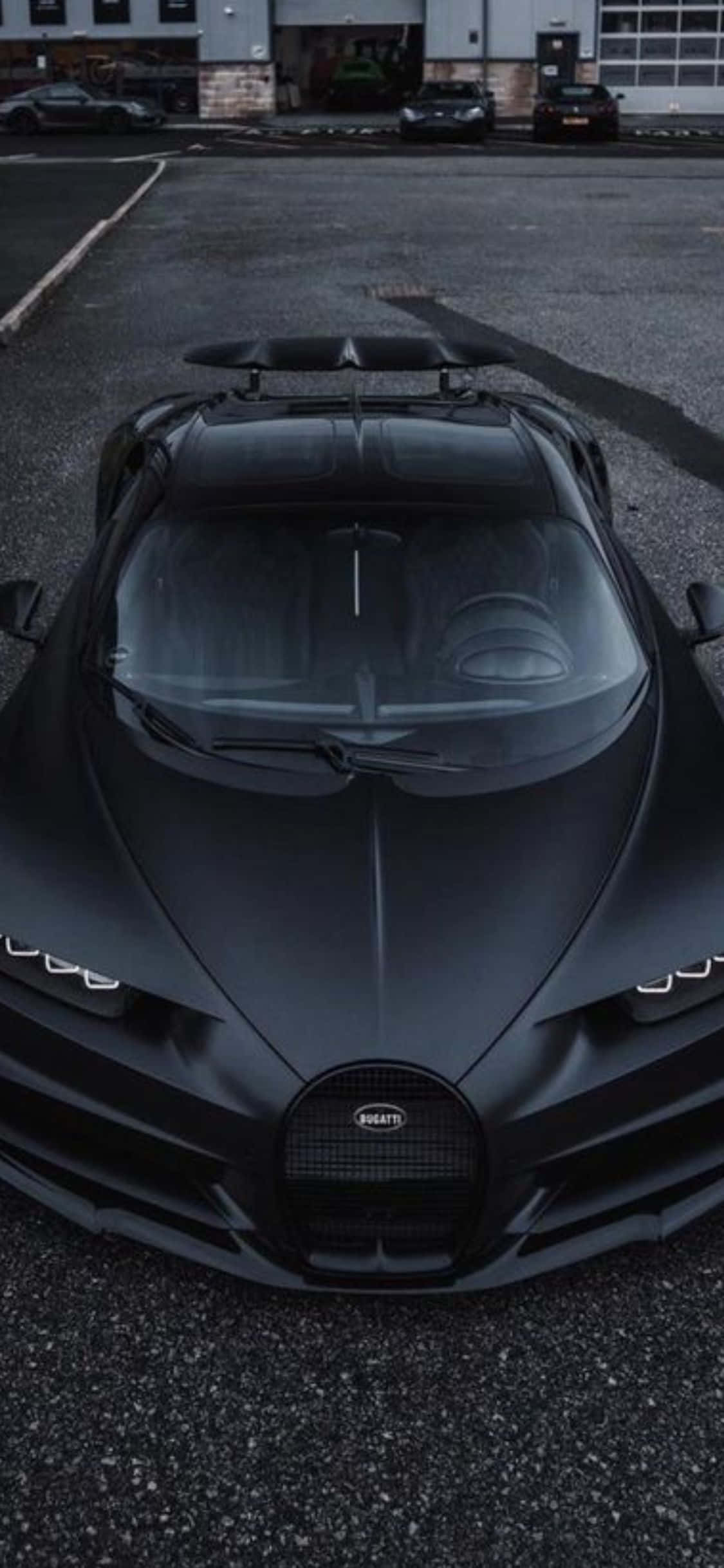 iPhone Xs Bugatti 桌布 壁紙 — 一輛消光黑Bugatti超跑在昏暗的瀝青停車場中，以流暢的空氣動力學線條和發光的車頭燈佔據主導地位。