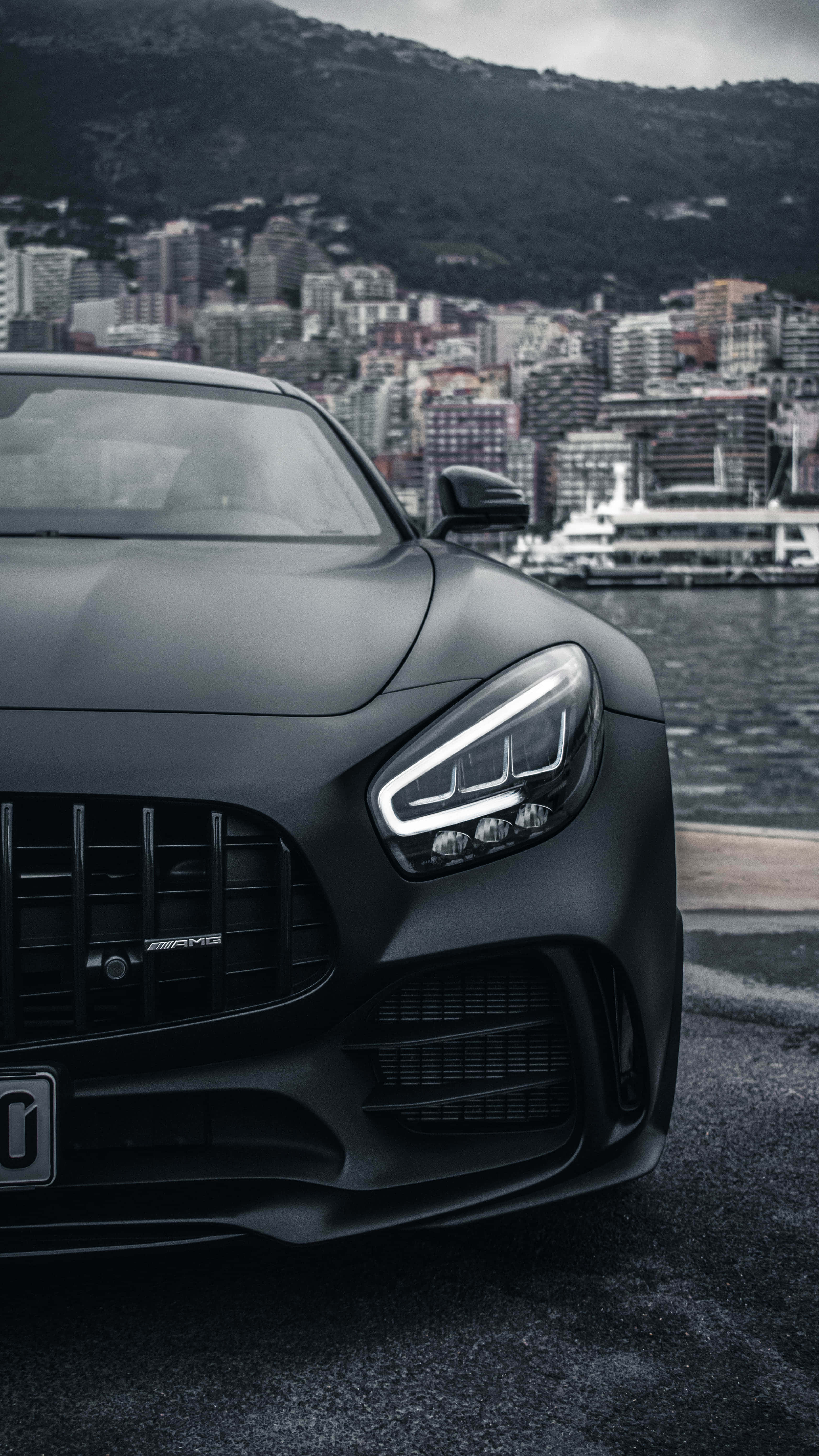 跑車 桌布 壁紙 — 一輛消光黑色 Mercedes-AMG 跑車停泊在海濱，背景是城市天際線和山脈。
