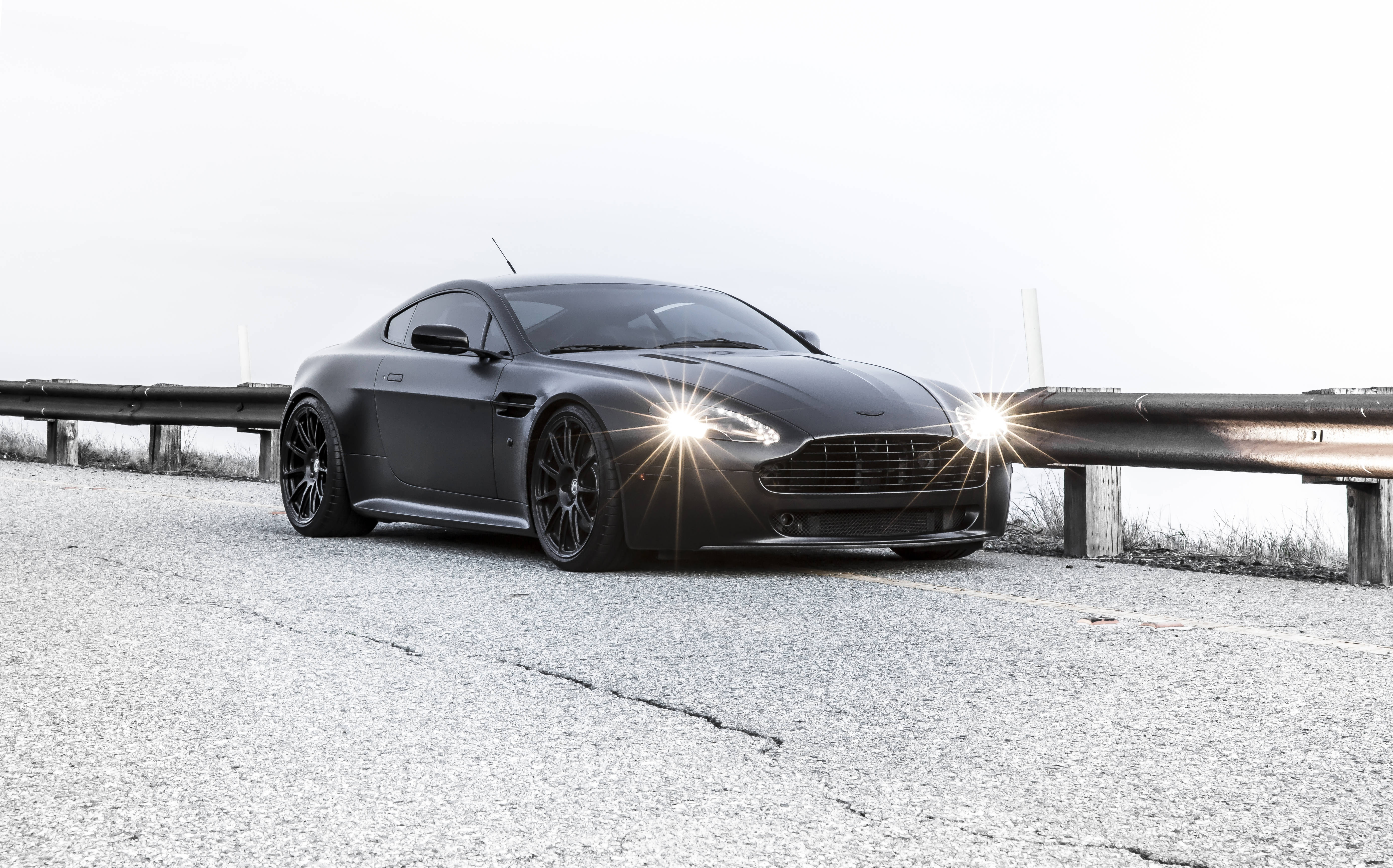 奧斯頓馬丁 桌布 壁紙 — 一輛消光黑色Aston Martin跑車，車頭燈發亮，停泊在裂開的柏油路上，旁邊有護欄，天空多雲。