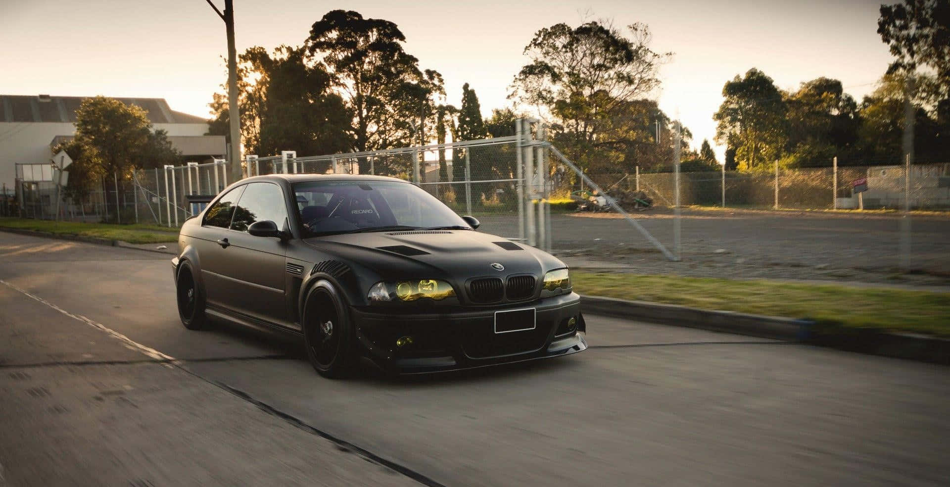 Tuned Car 桌布 壁紙 — 一輛消光黑色BMW M3雙門跑車在黃金時段沿著郊區街道疾駛，背景有樹木和圍籬。