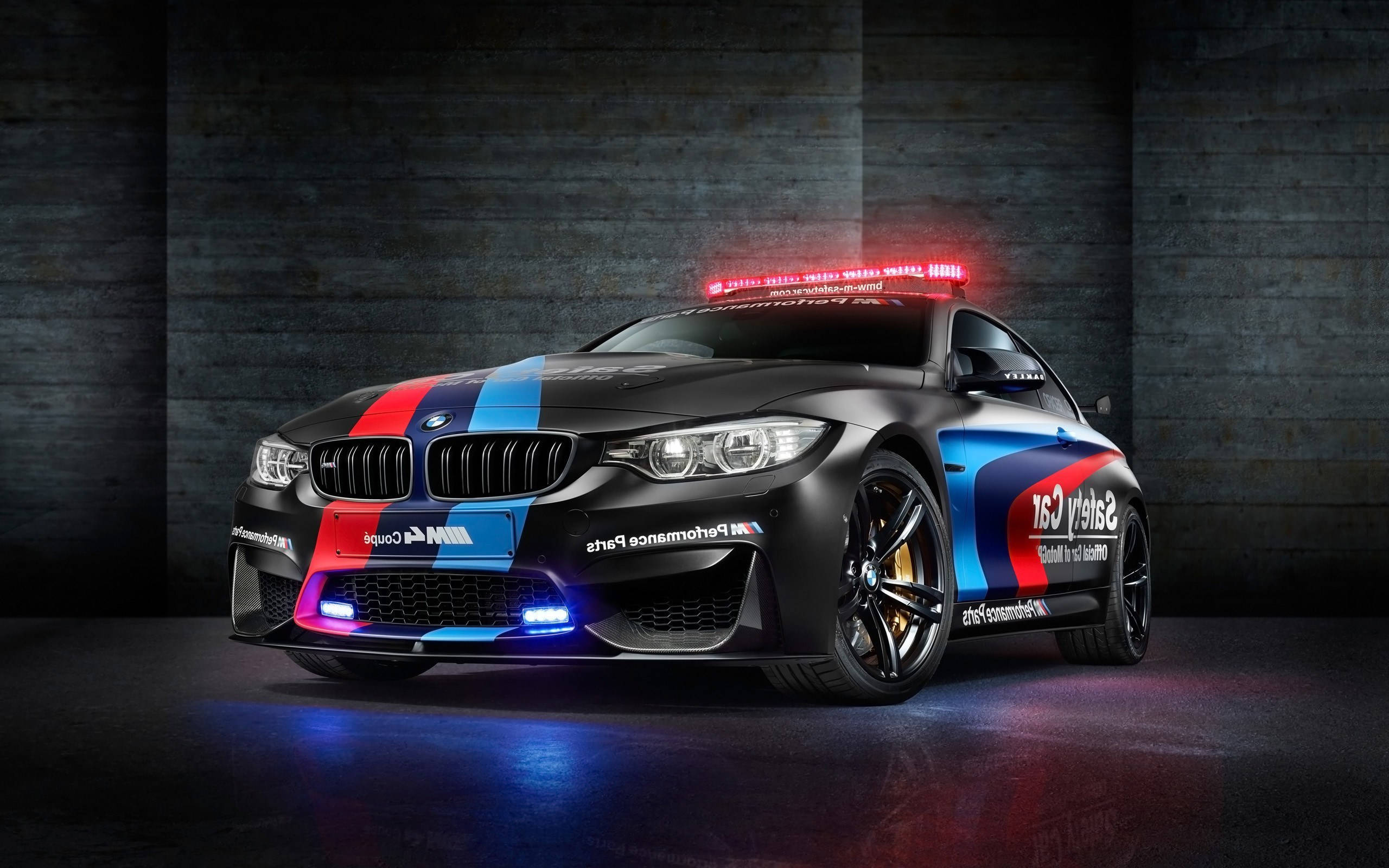 m 桌布 壁紙 — 一輛消光黑色BMW M4 Coupe安全車，帶有紅藍白賽車塗裝，在攝影棚燈光下於深色混凝土牆前閃耀。