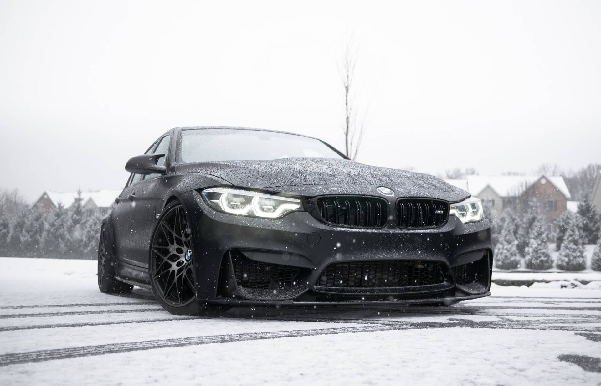 BMW 動圖 桌布 壁紙 — 一輛消光黑色BMW M4在郊區街道上飄移穿過飄落的雪花，背景是積雪的房屋和樹木。