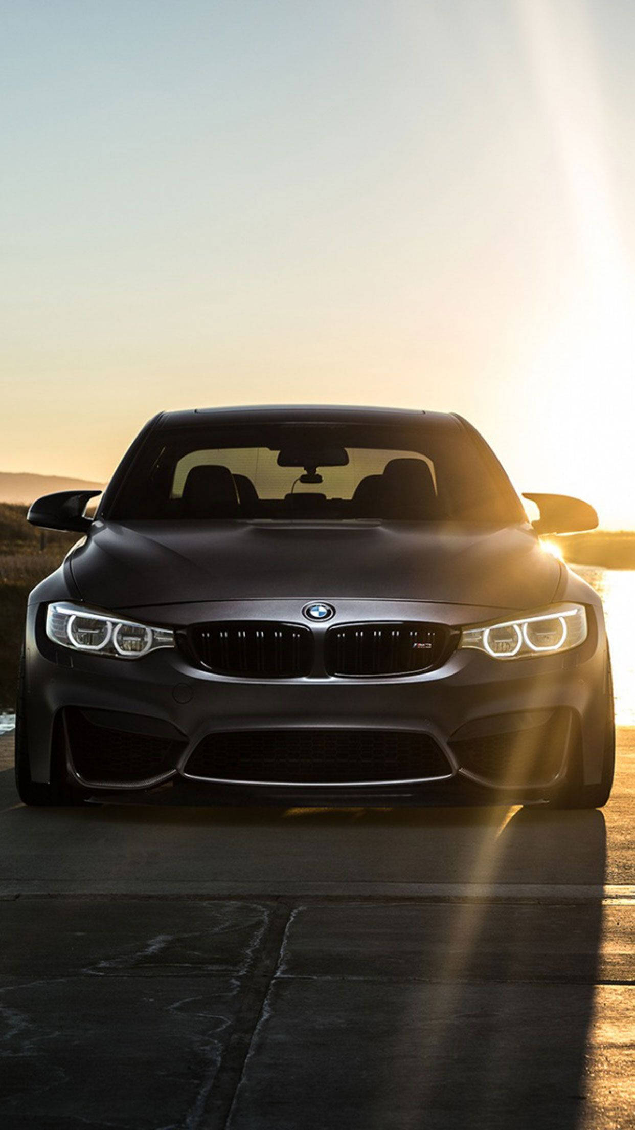 X 桌布 壁紙 — 一輛消光黑色BMW M4正面朝前，車頭燈發光，映襯著金色的日落天空。