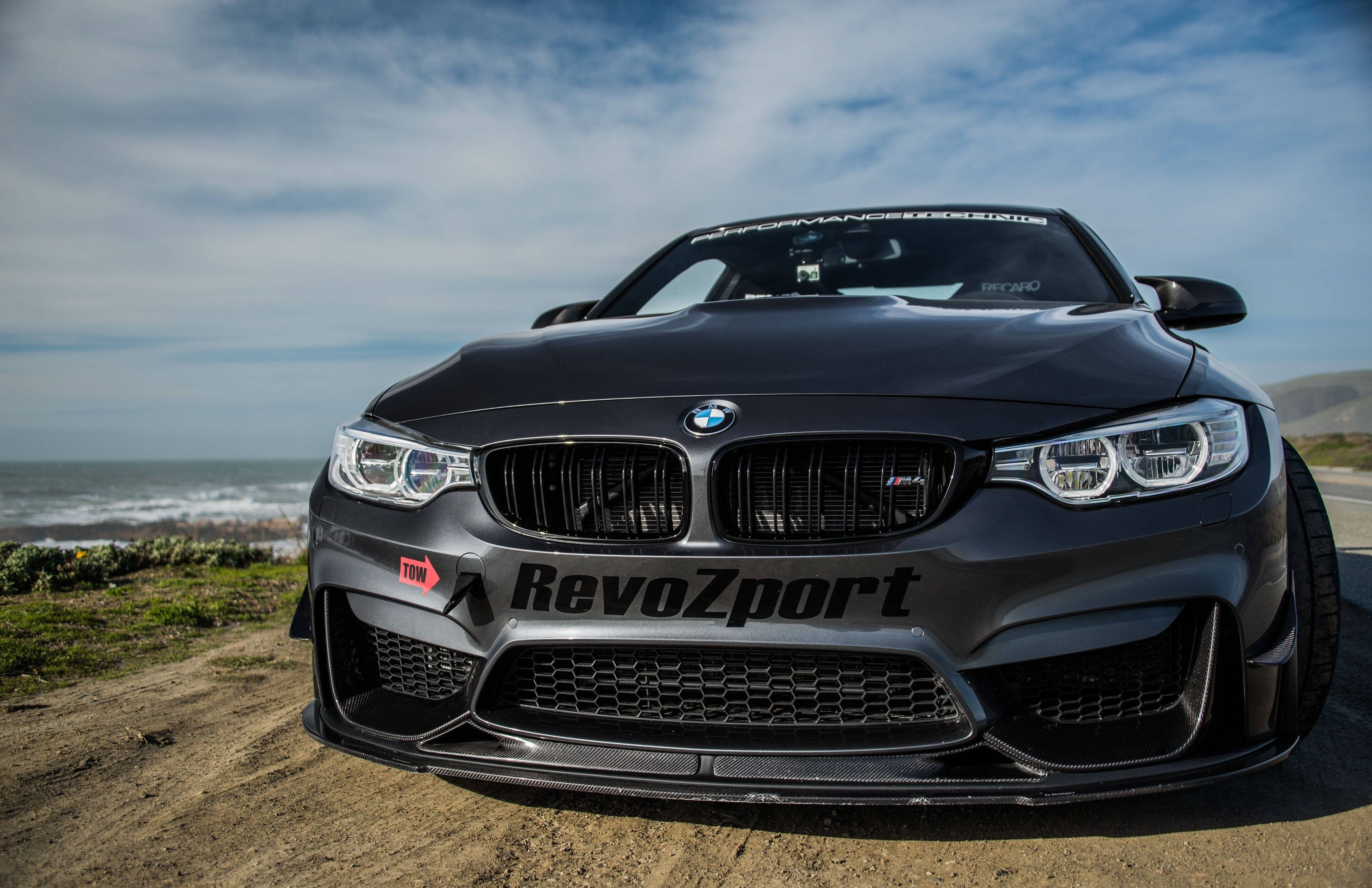 4K BMW 壁紙 桌布 壁紙 — 一輛消光黑色BMW M4配備RevoZport調校，在部分多雲的天空下停泊在沿海泥路上。