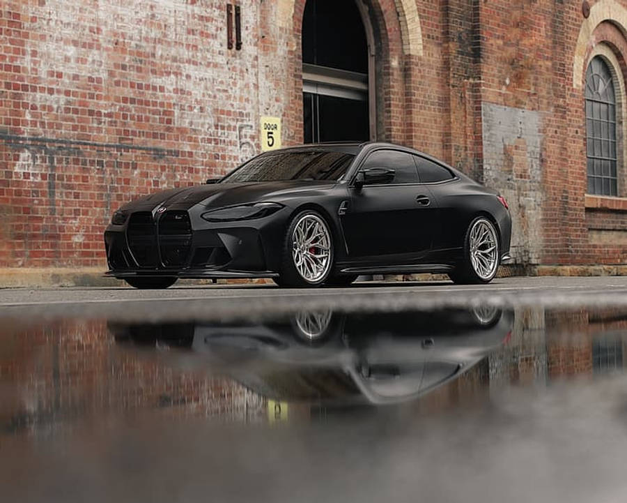 Bmw M4 桌布 壁紙 — 一輛消光黑色BMW M4雙門跑車在潮濕的路面上反射，旁邊是帶有拱形窗戶的風化磚倉庫。