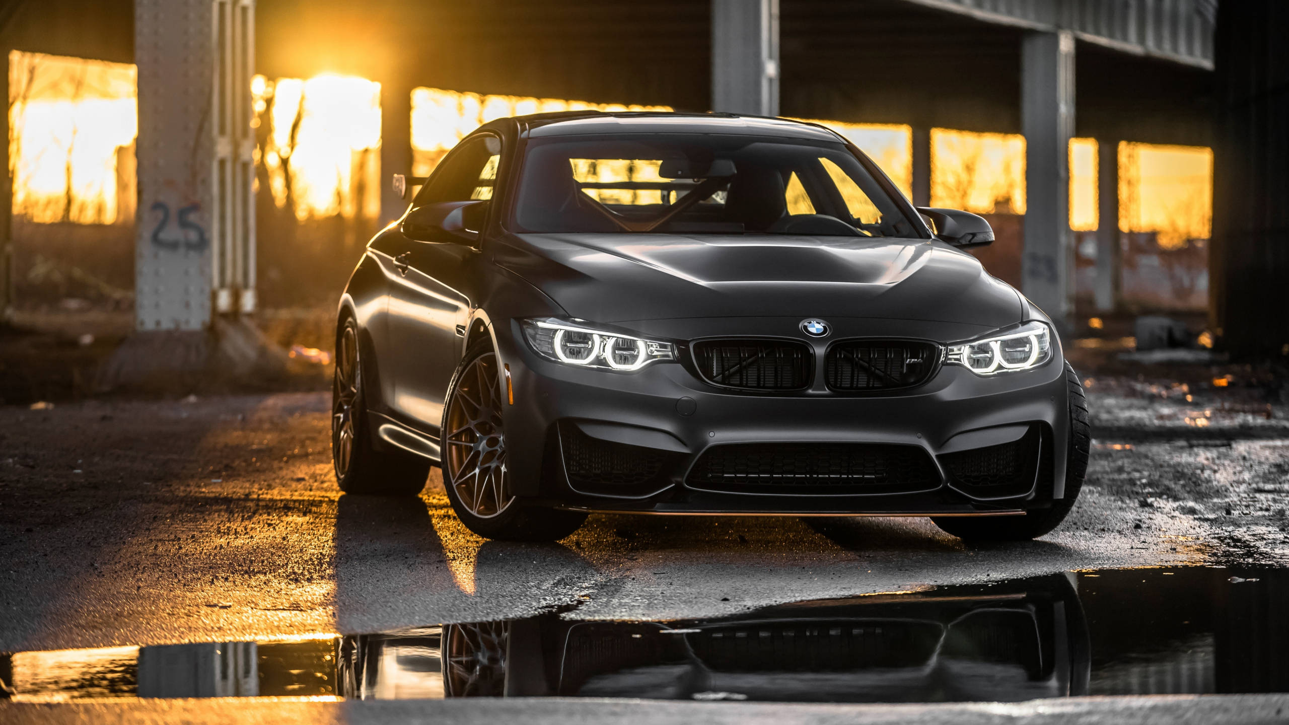 BMW 動圖 桌布 壁紙 — 一輛消光黑色BMW M4雙門跑車在金色夕陽下閃耀，其倒影在工業結構下的水坑中閃爍。