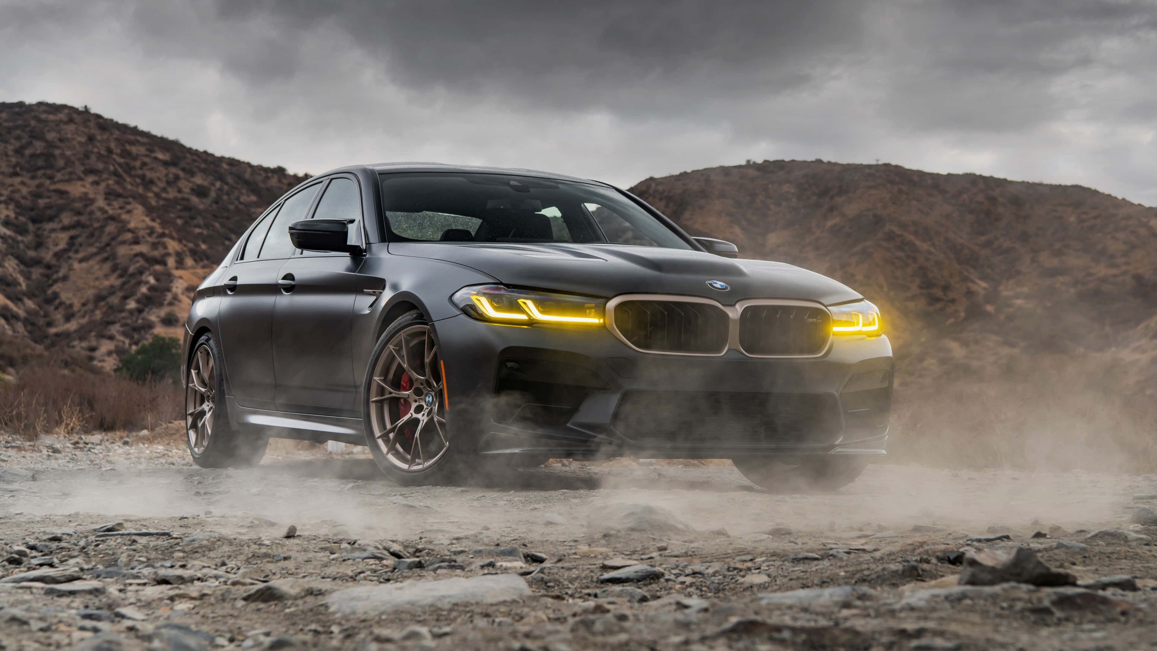 桌布 壁紙 — 一輛消光黑色BMW M5在塵土飛揚的地形中飄移，黃色車頭燈在暴風雨的天空下閃耀。