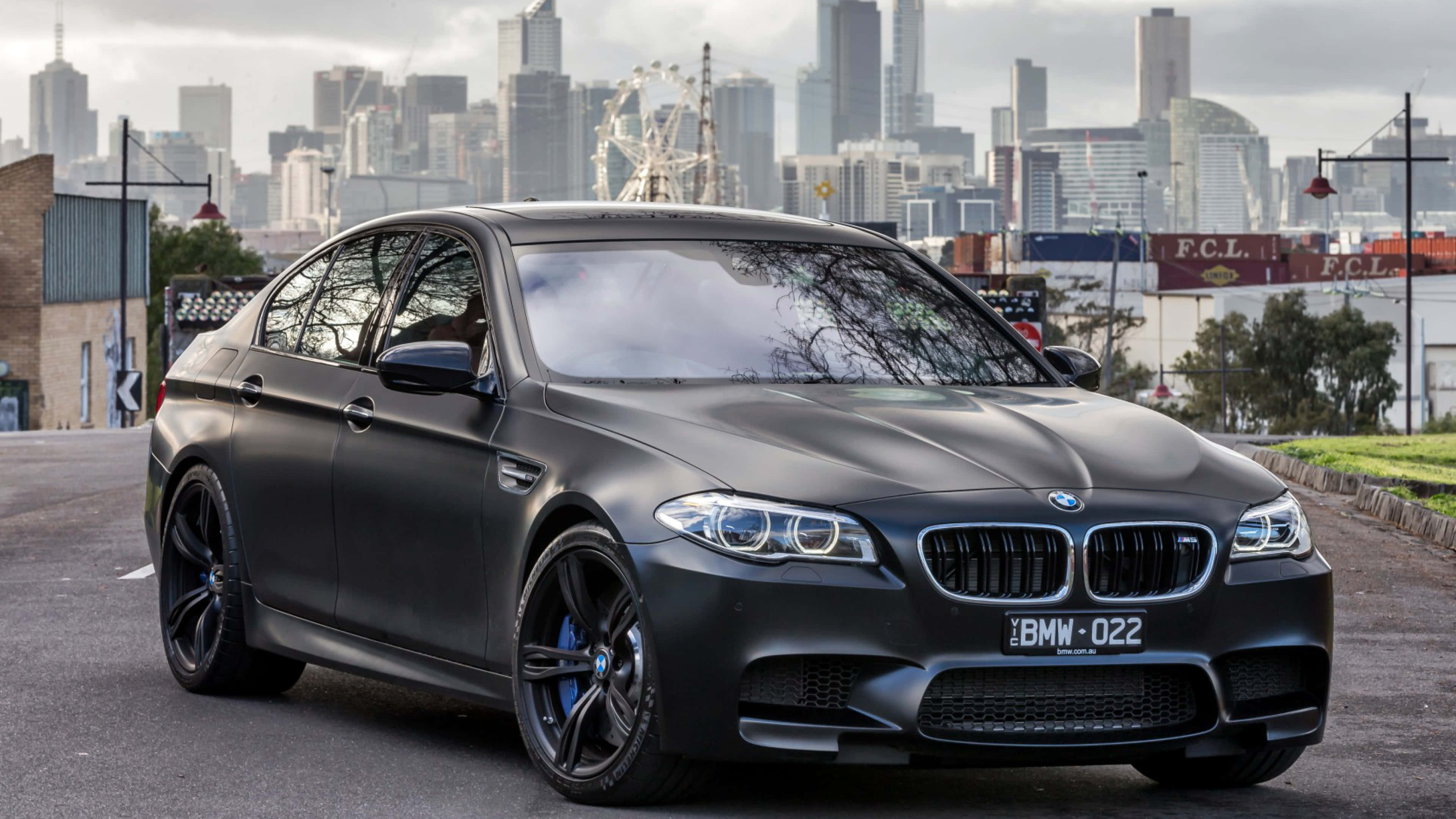 桌布 壁紙 — 一輛消光黑色BMW M5跑車停放在城市街道上，背景是現代天際線和摩天輪。