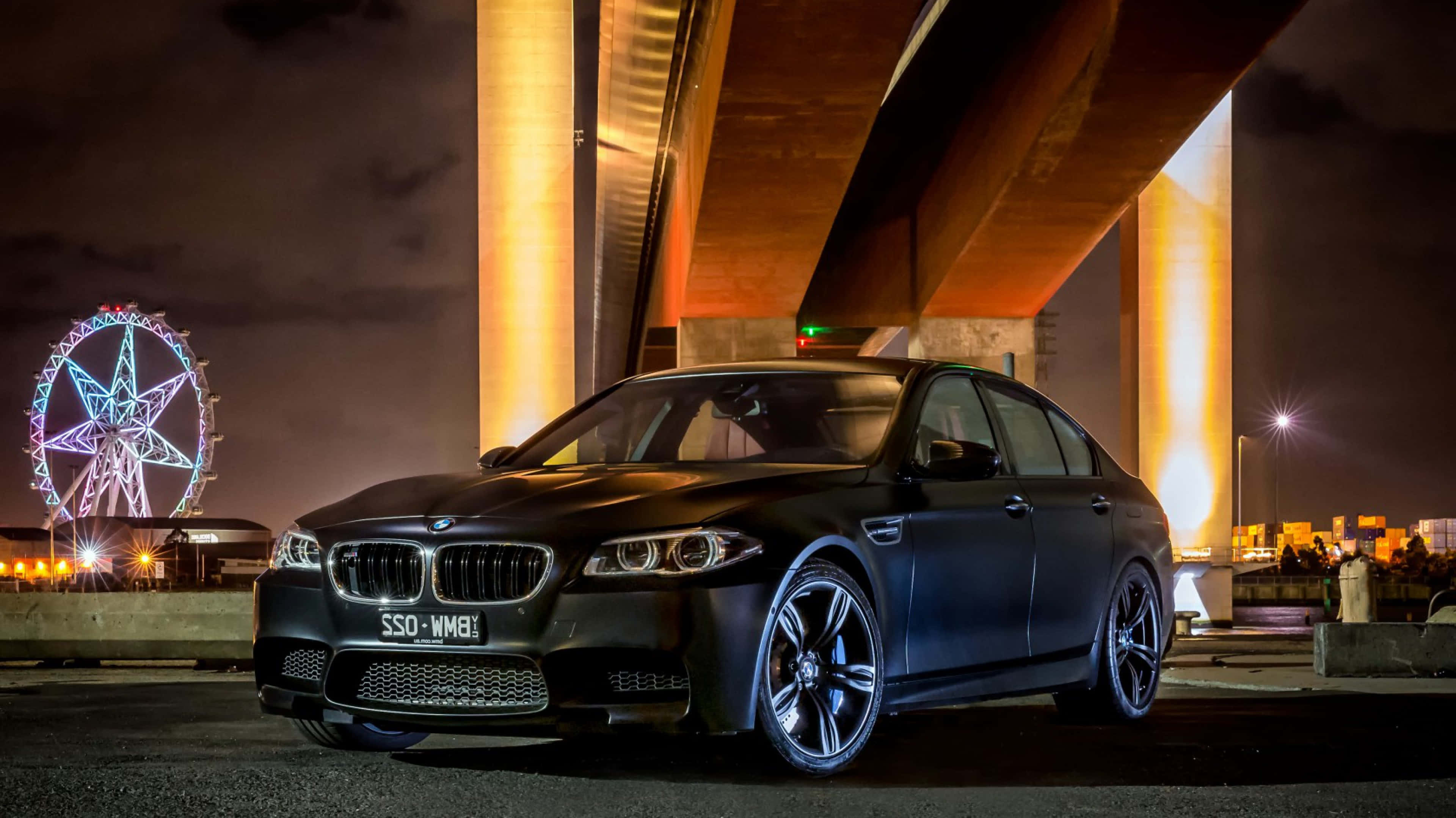 桌布 壁紙 — 一輛消光黑色BMW M5轎車在夜間停放在發光的橋下，背景是色彩繽紛的摩天輪。
