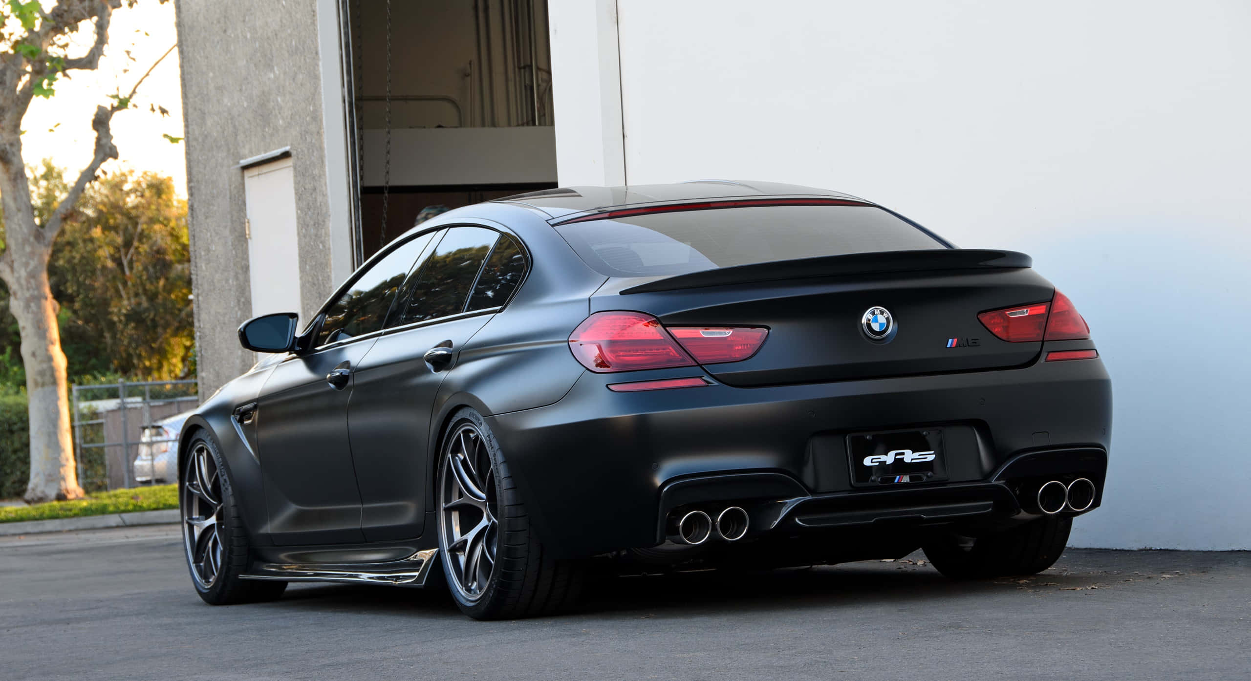 Bmw M6 桌布 壁紙 — 一輛消光黑色BMW M6 Gran Coupe，配備四出排氣管和紅色尾燈，在黃金時段停放在混凝土建築物旁。