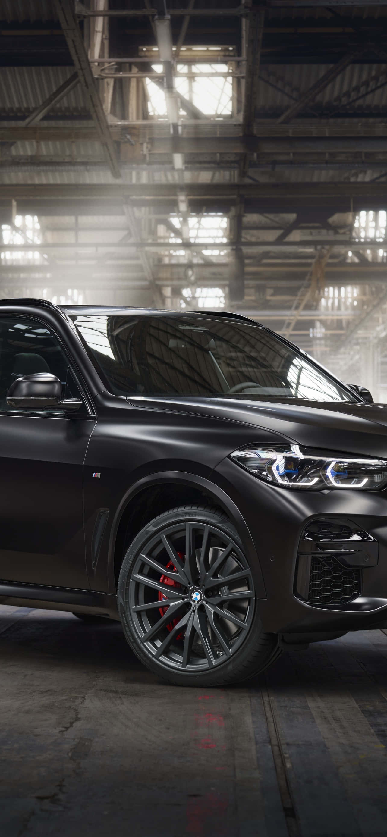 Bmw X6 M 桌布 壁紙 — 一輛消光黑色BMW SUV，帶紅色煞車卡鉗，停放在一個灰塵滿佈、高天花板且陽光穿透的工業倉庫中。