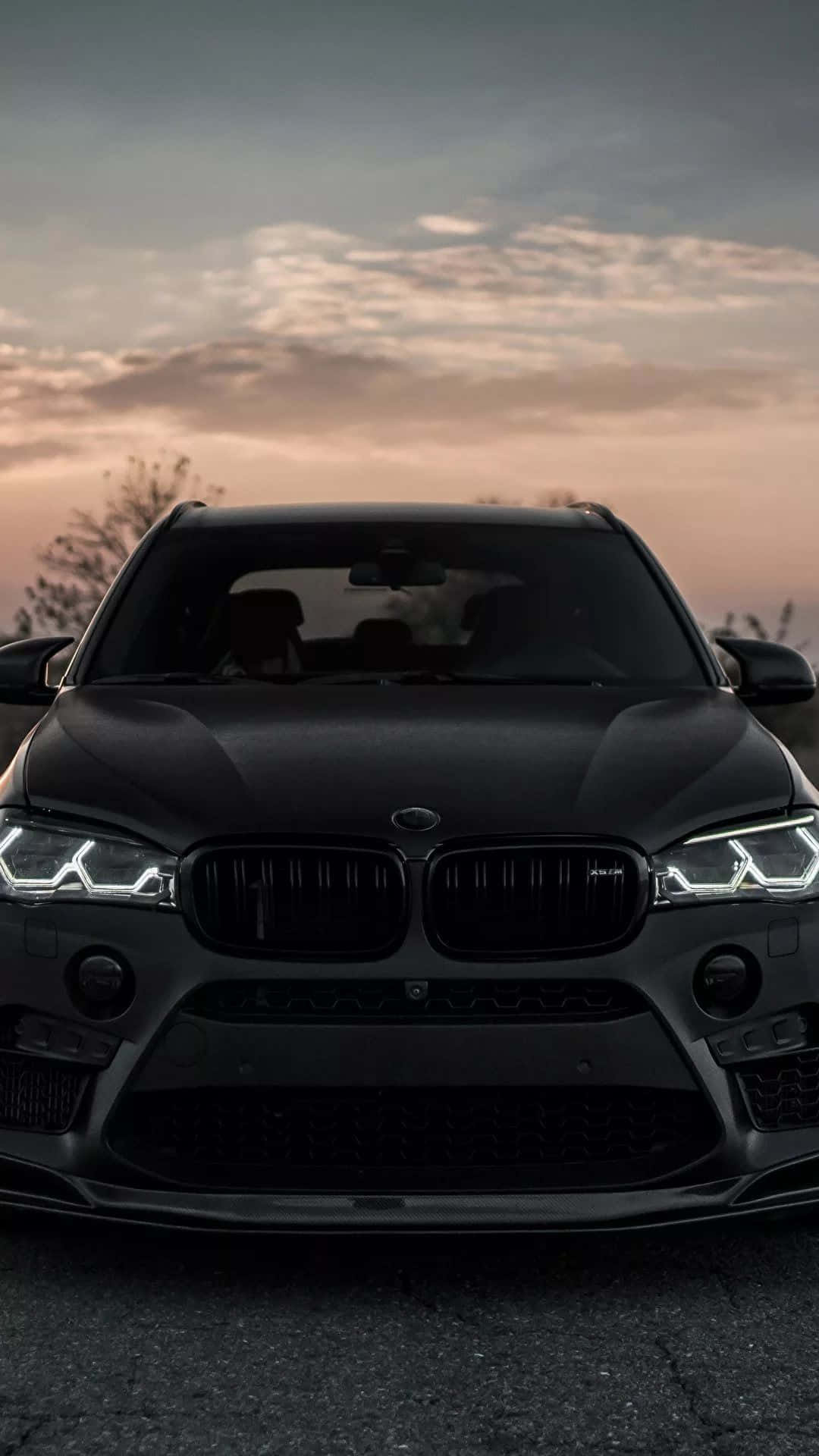 BMW M 桌布 壁紙 — 一輛消光黑色BMW SUV正面向前，車頭燈發光，背景是柔和粉紅與橙色的日落天空。
