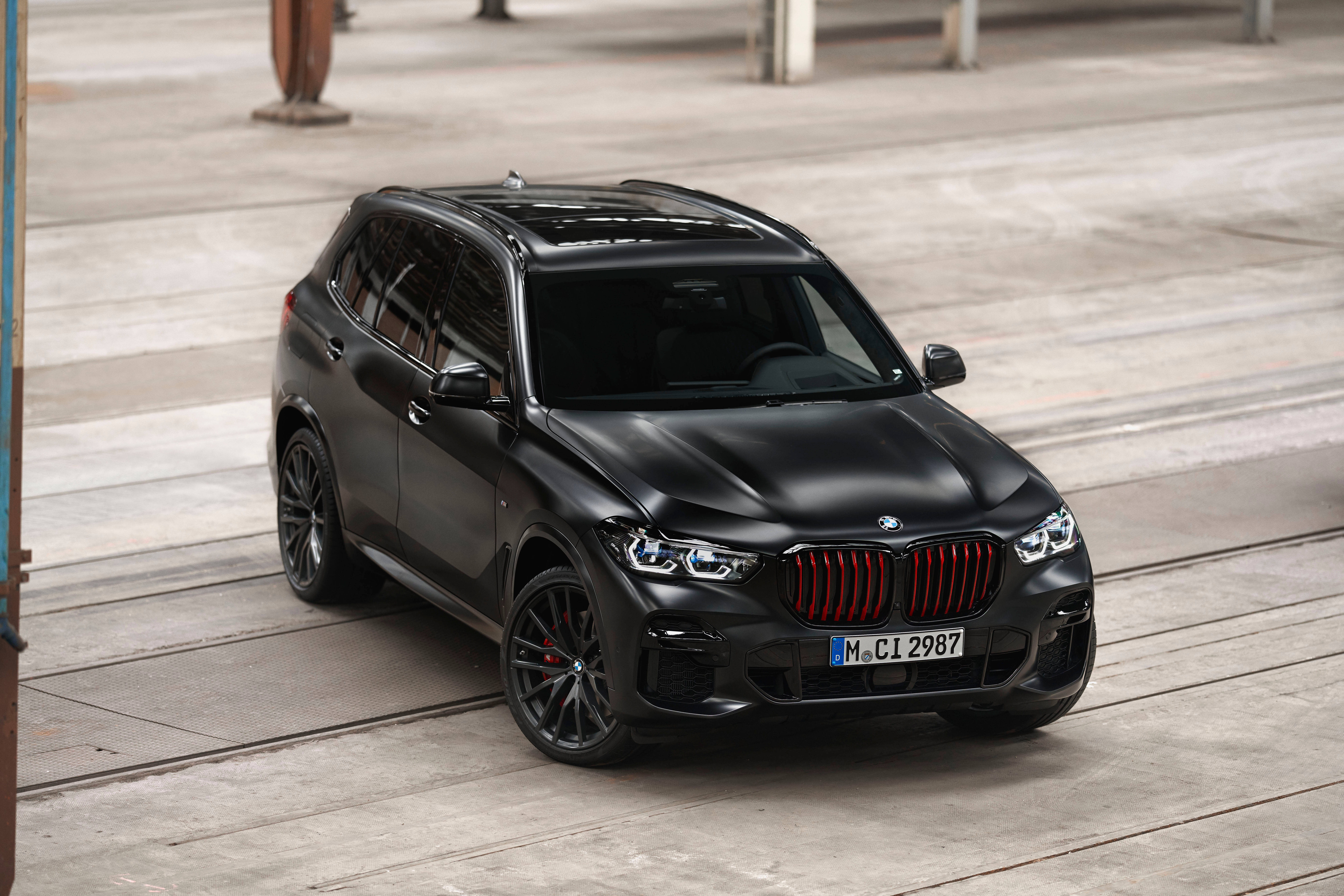 Black Bmw 桌布 壁紙 — 一輛消光黑色BMW X5 M Competition SUV，帶紅色飾件，停放在工業橫樑下的混凝土上。