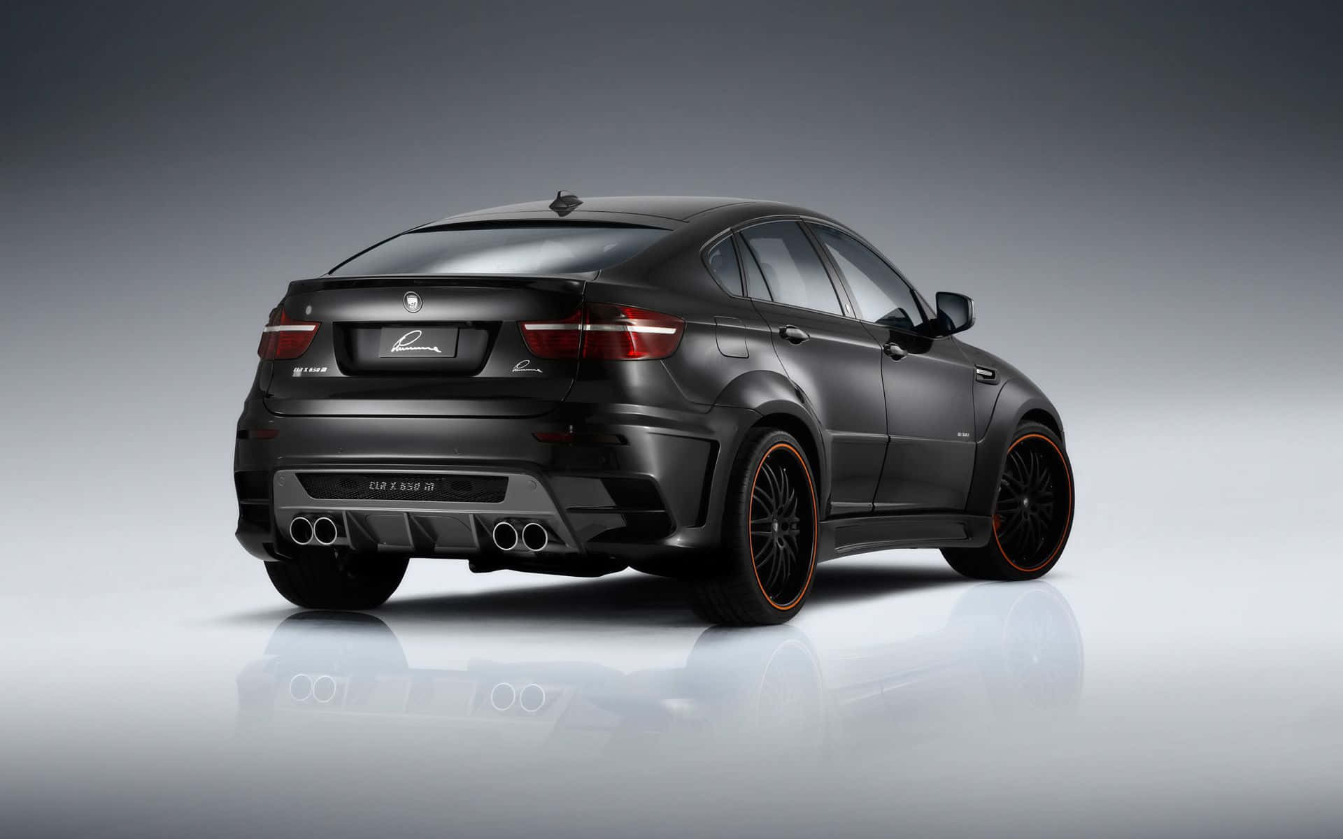Bmw X6 桌布 壁紙 — 一輛消光黑色BMW X6M，配備橙色輪圈和四出排氣管，在反射地板的攝影棚中展示。
