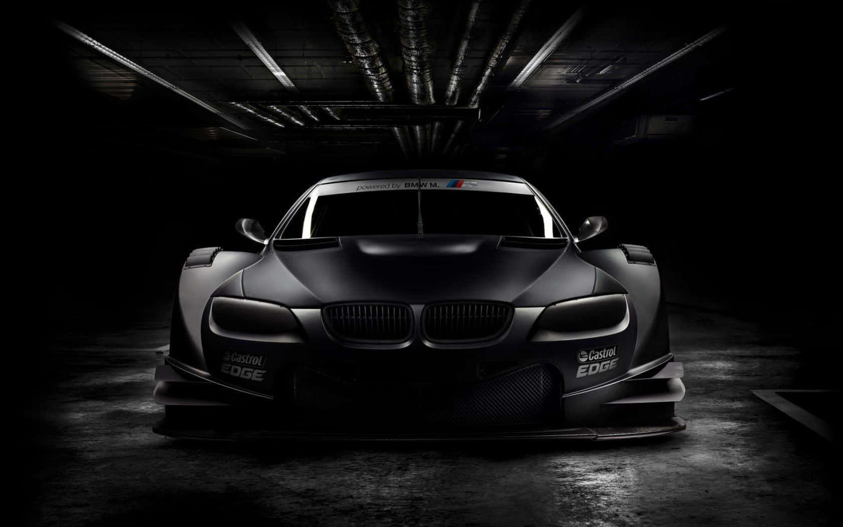 Black And White Car 桌布 壁紙 — 一輛消光黑色BMW賽車在昏暗的工業車庫中，在戲劇性的燈光下向前凝視。